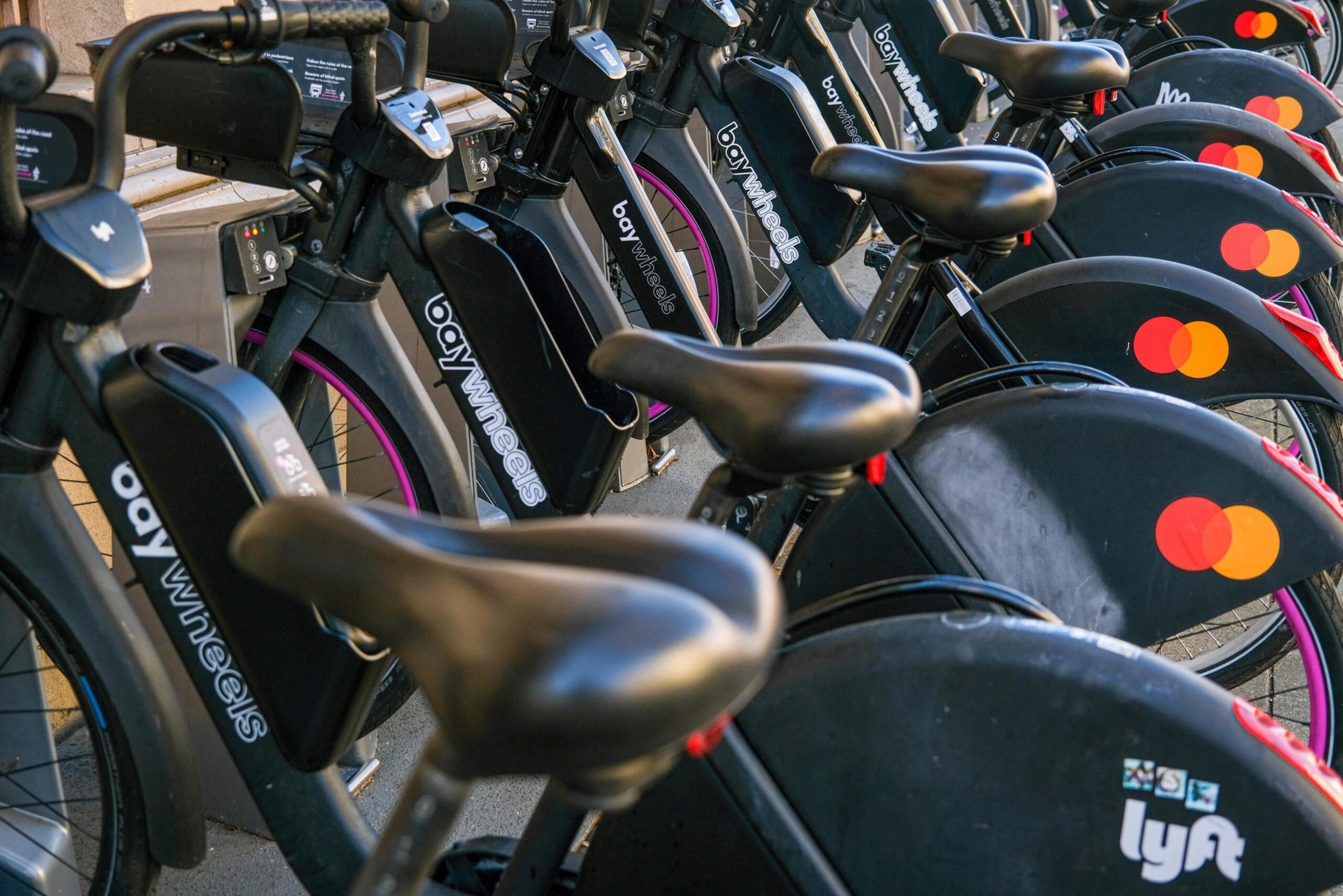 A row of black Bay Wheels bikes with purple rims and black seats are parked closely together on a sidewalk.