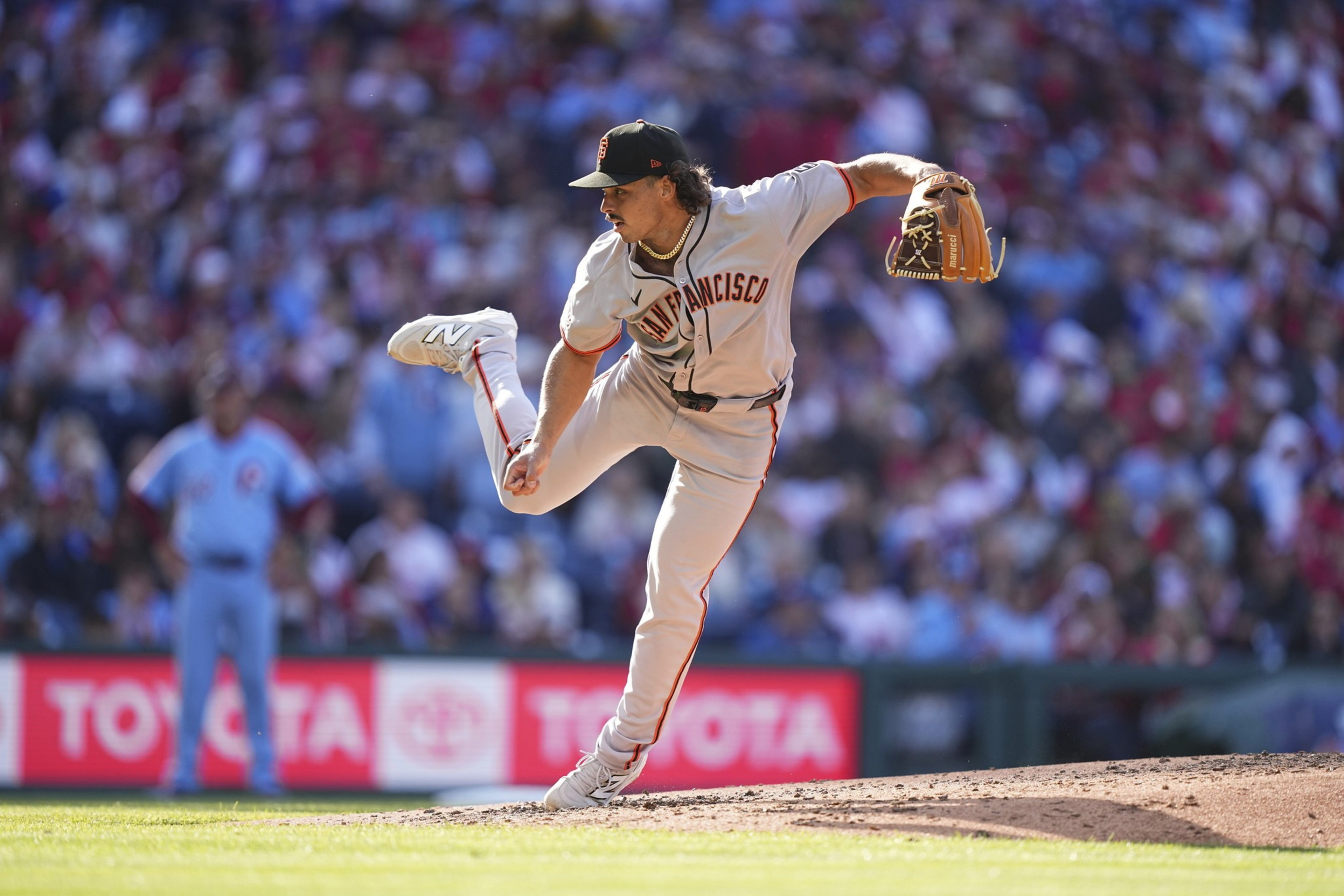 A baseball pitcher in a "San Francisco" uniform is mid-throw on the mound, with one leg raised high. A blurred crowd and field advertisements are in the background.