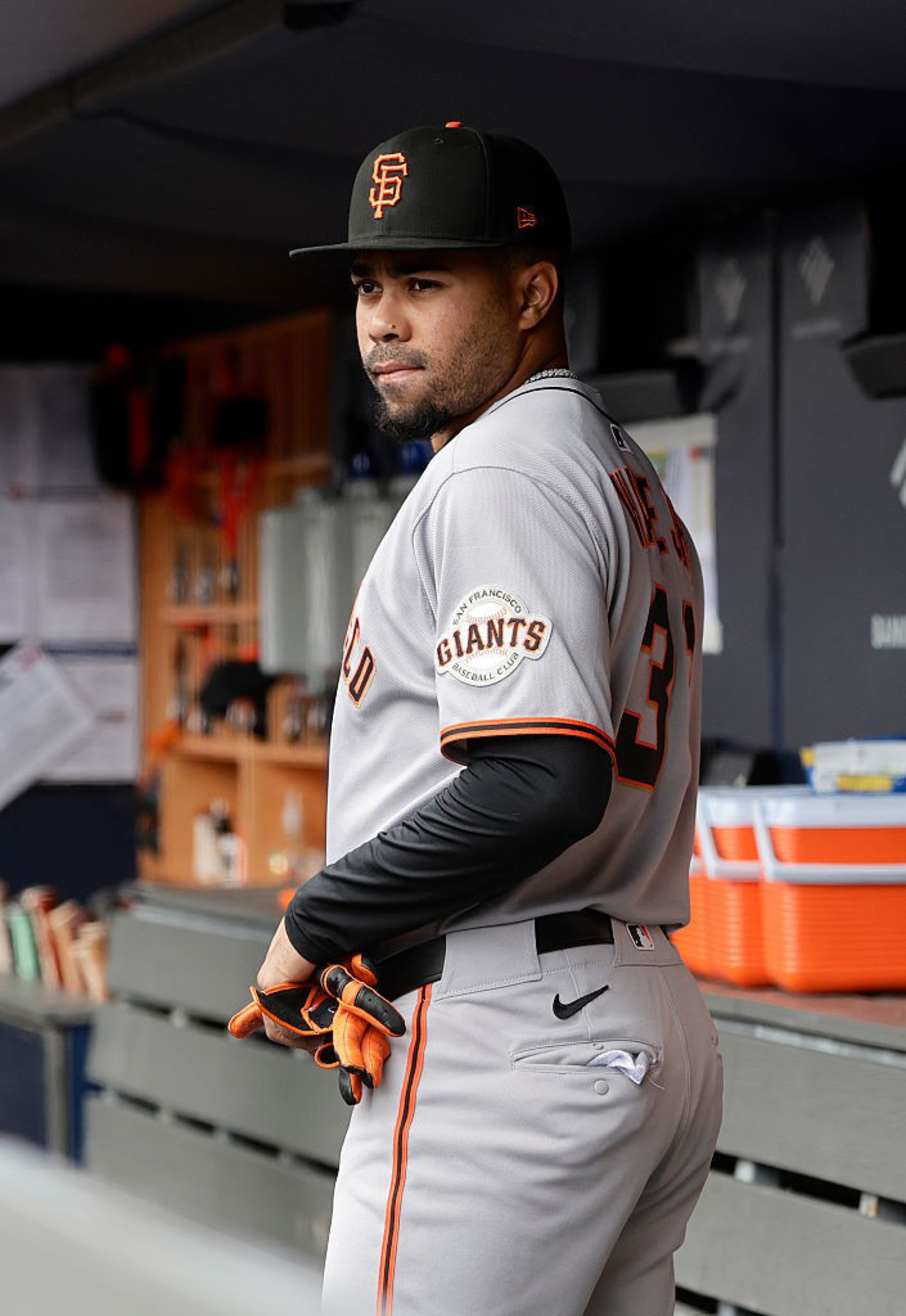 A baseball player in a gray uniform and black cap stands in a dugout, wearing gloves and facing sideways. The uniform has "Giants" written on it.