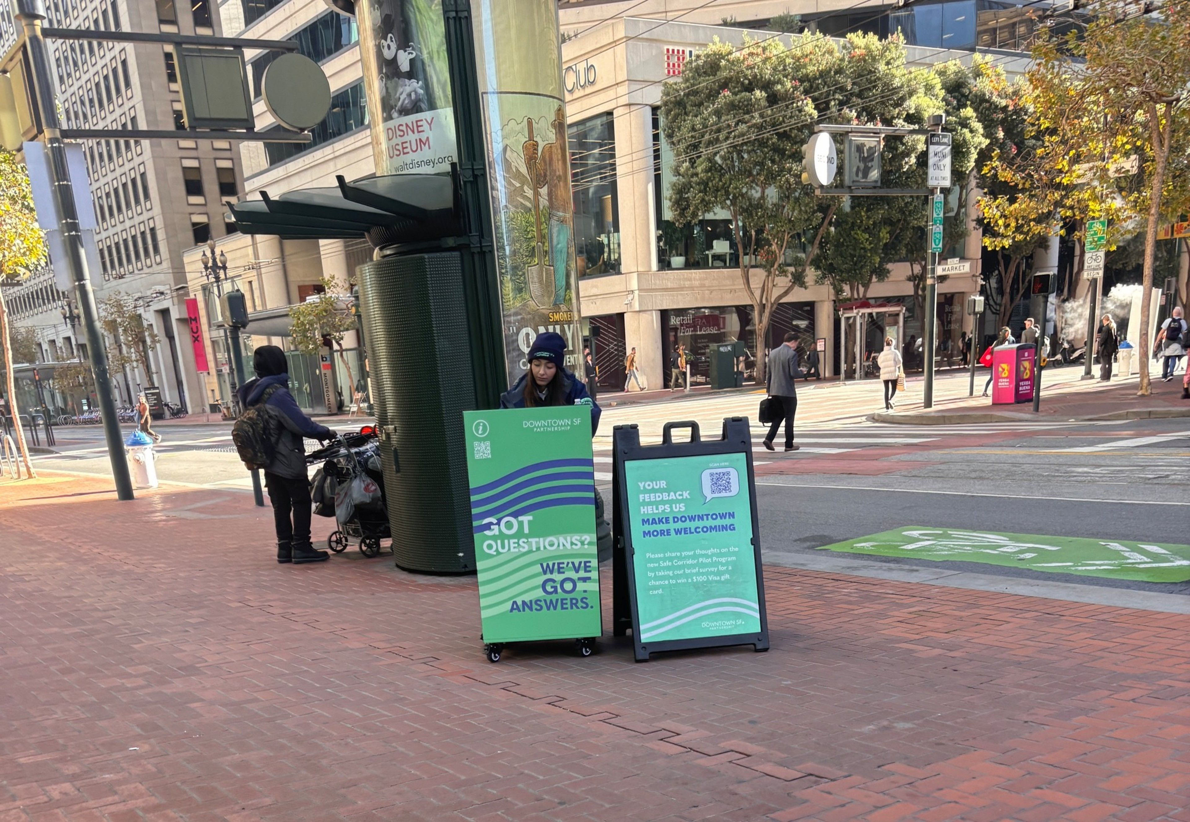 A person stands behind two green signs on a brick sidewalk in an urban area, with buildings, trees, and pedestrians visible in the background.