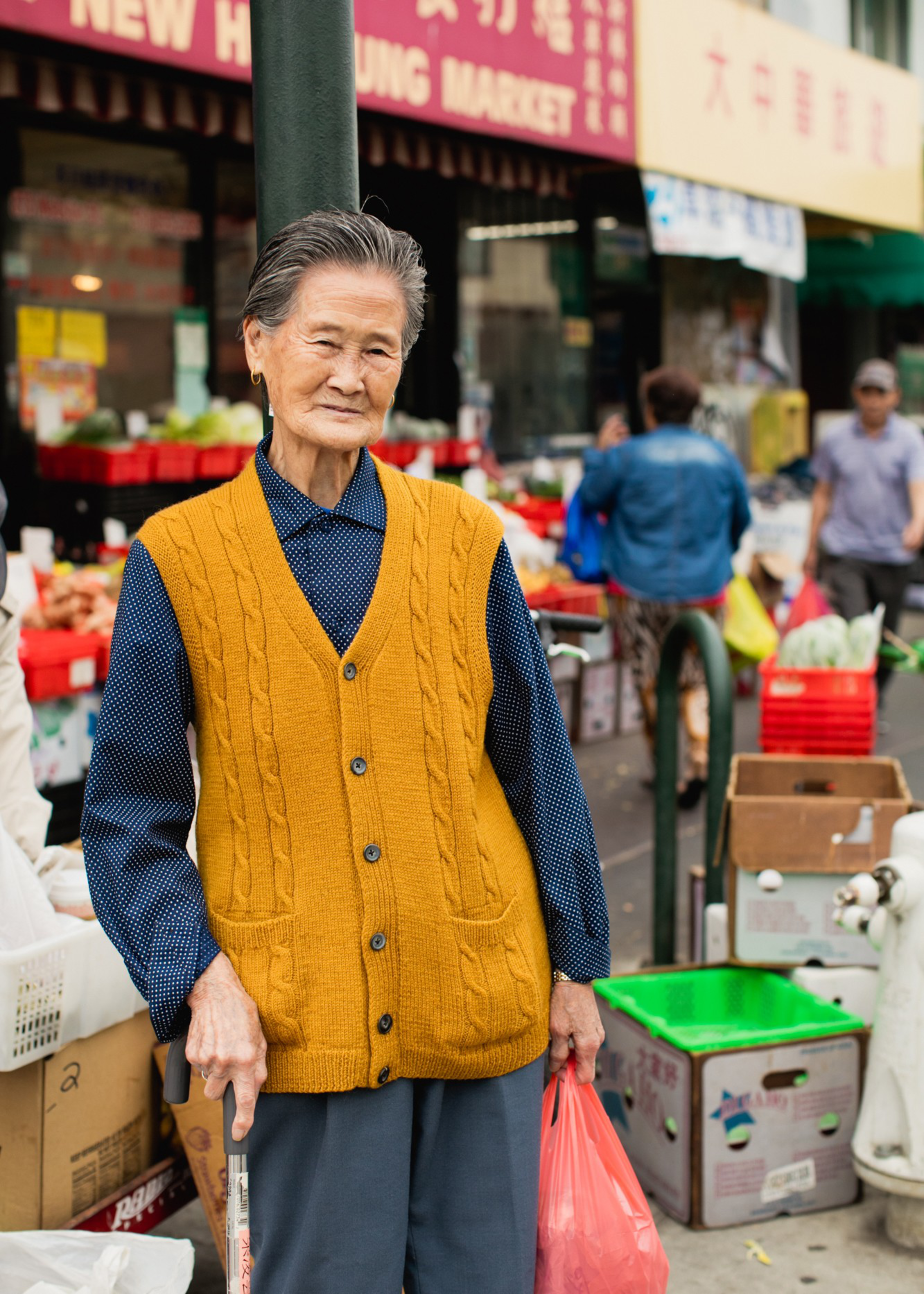An elderly woman wearing a mustard yellow vest and blue shirt holds a red plastic bag and a walking cane, standing in a busy outdoor market.