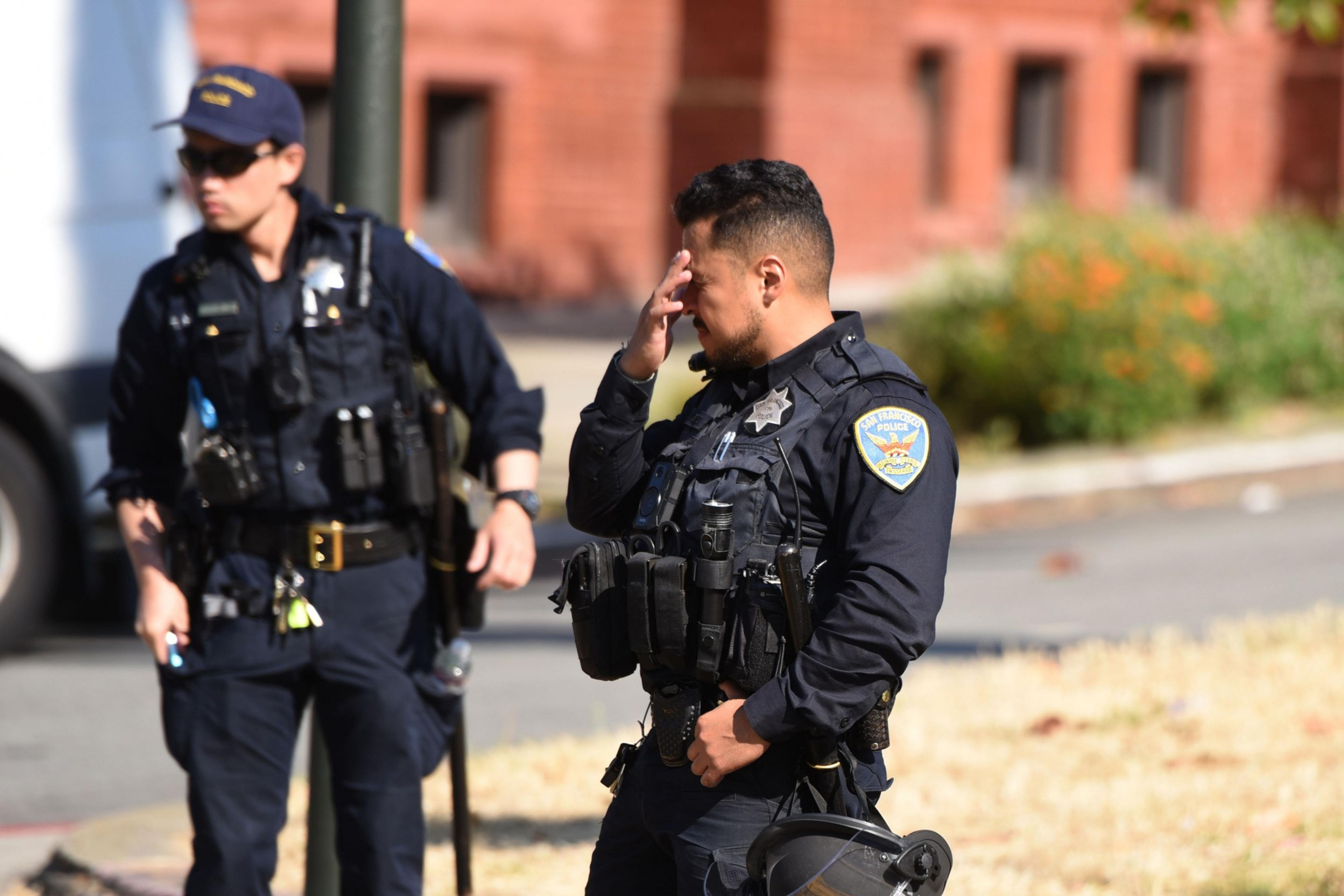 Two police officers in uniforms stand outdoors. One holds a helmet and wipes his forehead, while the other stands nearby, holding a baton. Buildings and greenery are in the background.