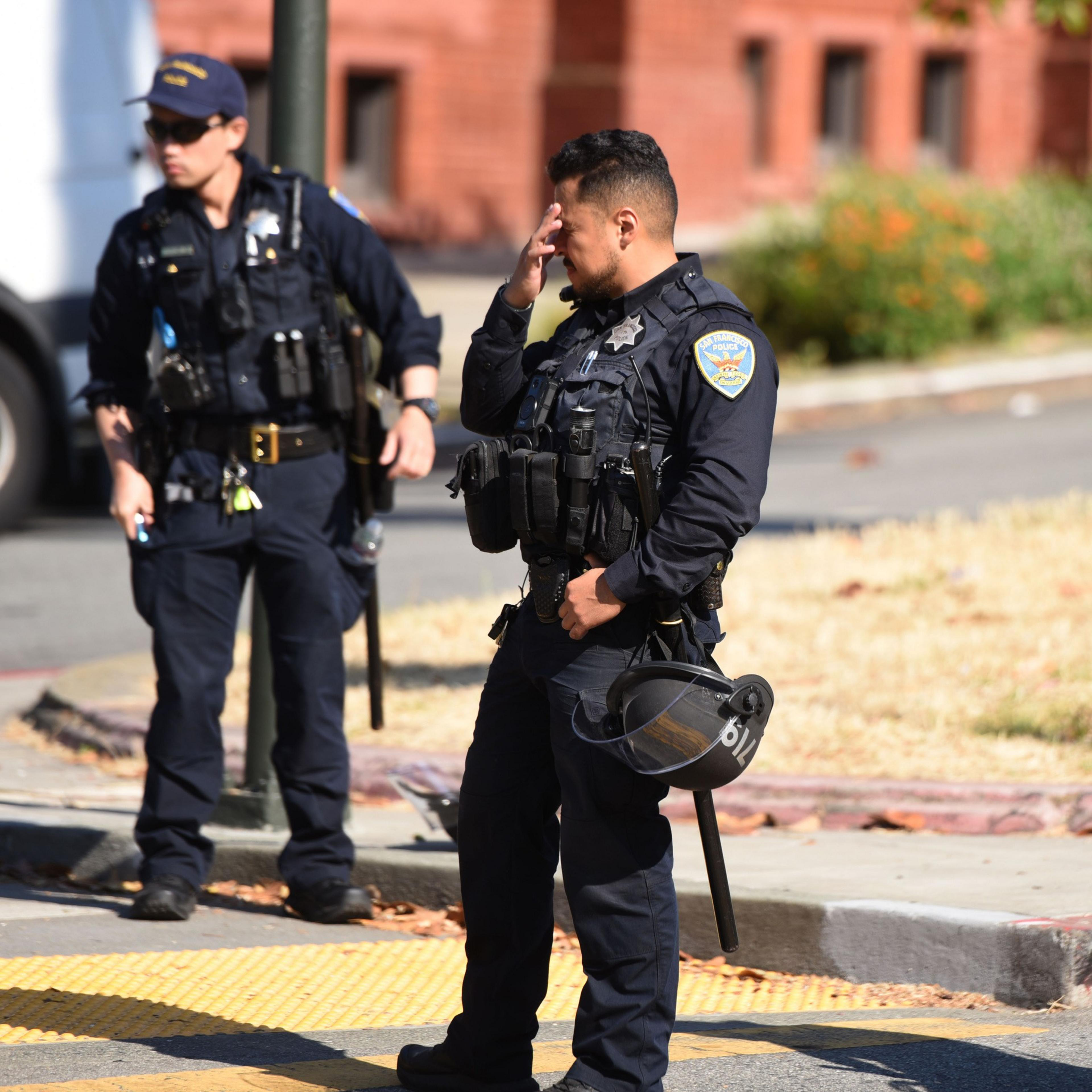 Two police officers in uniforms stand outdoors. One holds a helmet and wipes his forehead, while the other stands nearby, holding a baton. Buildings and greenery are in the background.