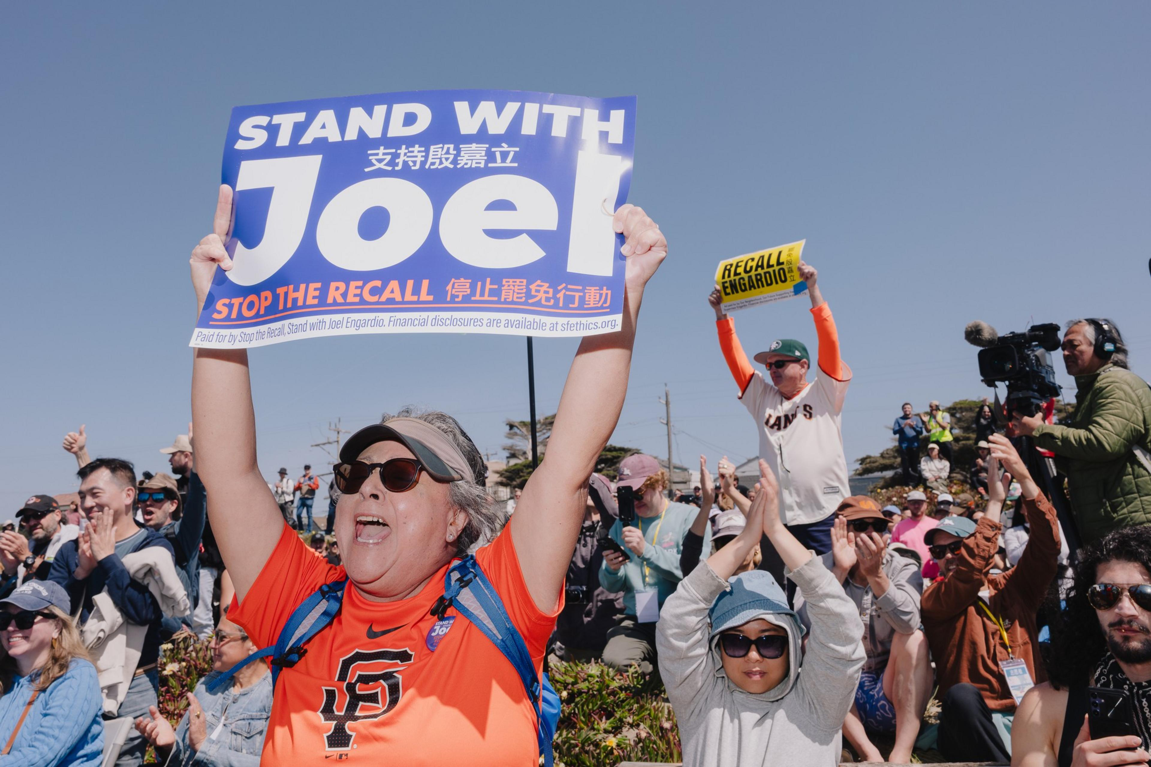 A crowd gathers, with people holding signs. A woman in front holds a "Stand with Joel" sign, while another sign says "Recall Engardio." The crowd appears engaged.