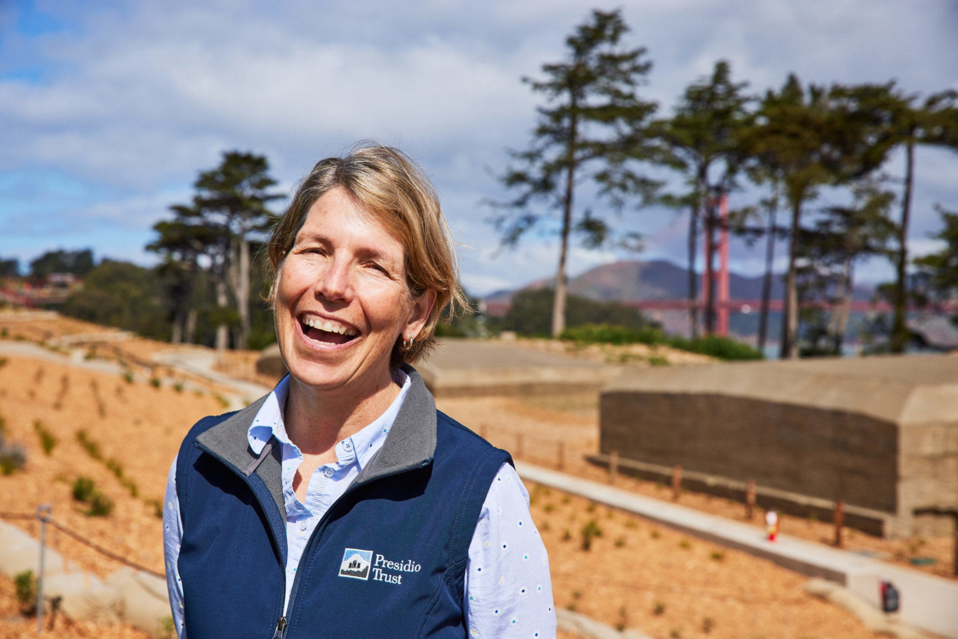 A smiling woman wearing a blue Presidio Trust vest stands outdoors with trees, a walkway, and the Golden Gate Bridge faintly visible in the background.