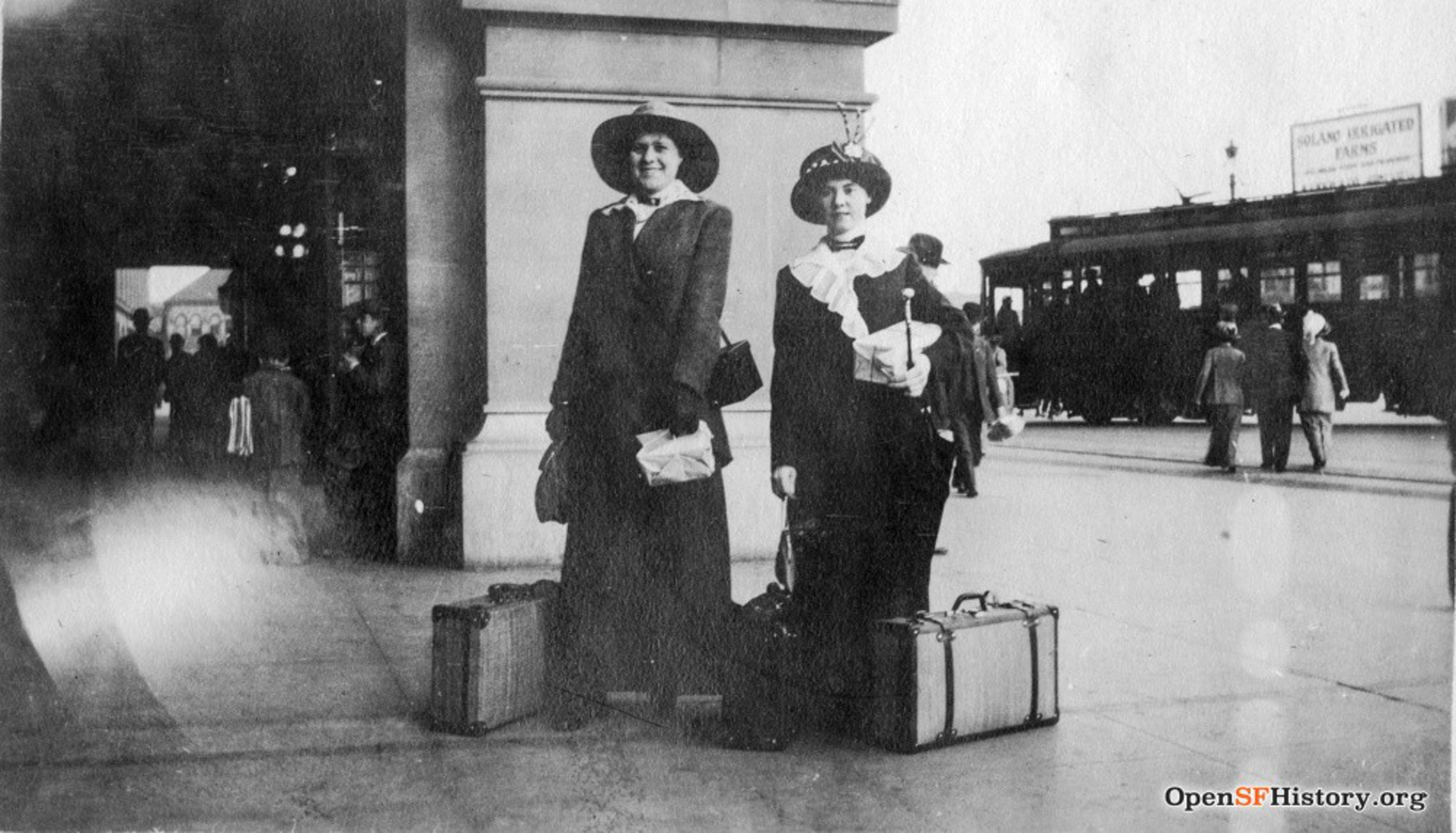 Two women in early 20th-century attire stand with luggage at a busy streetcorner with pedestrians and a streetcar in the background.