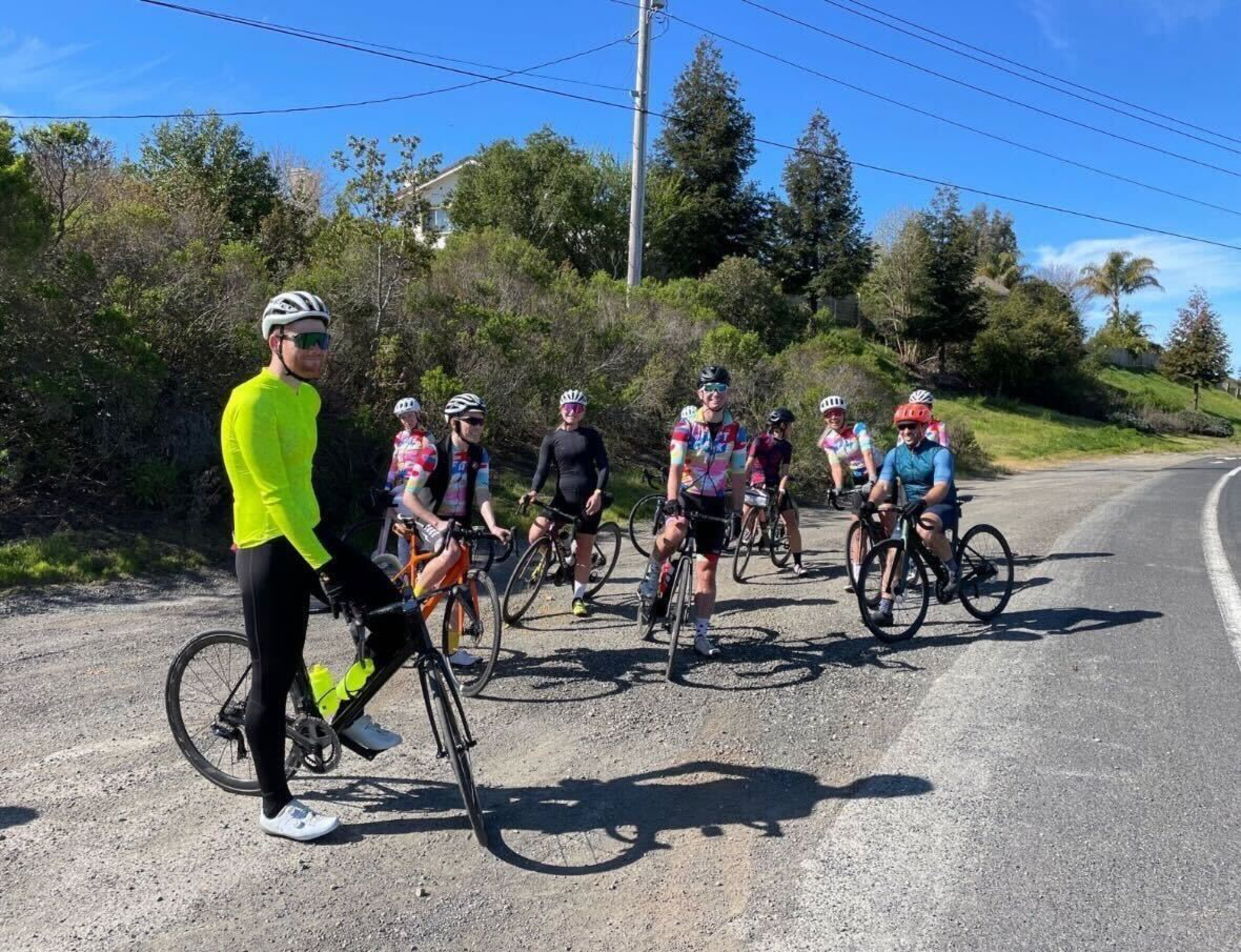 A group of eight cyclists in helmets and colorful gear pauses with their bikes on a roadside gravel patch under a clear blue sky.