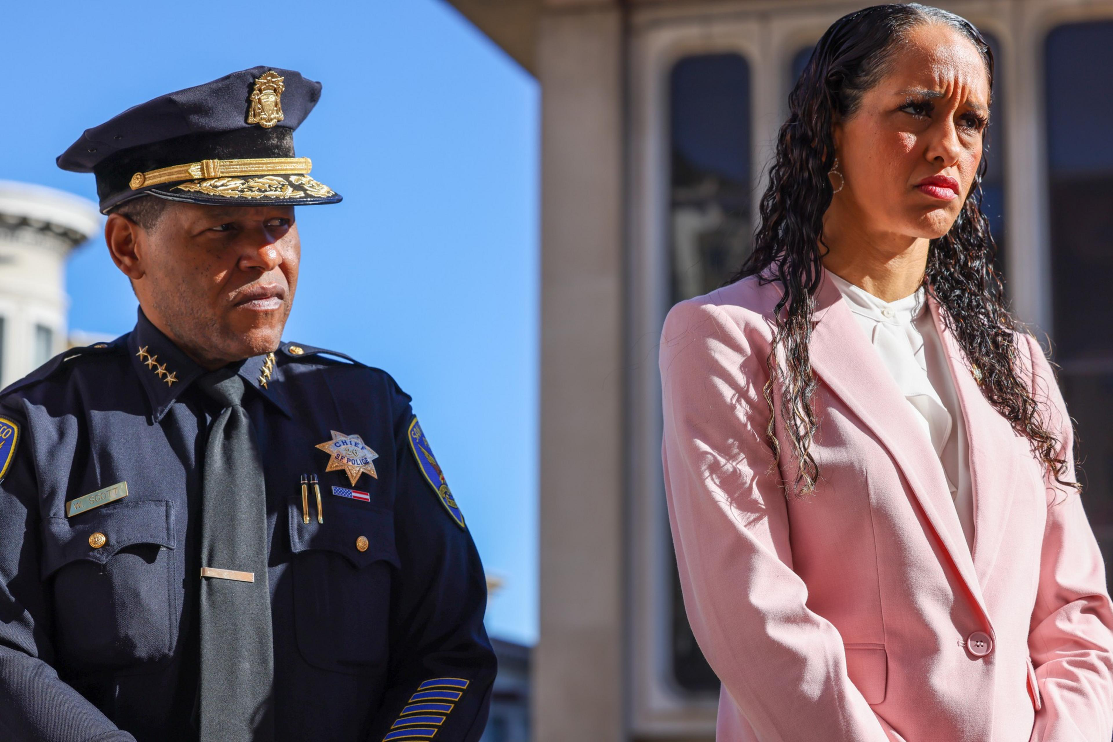 A police officer in uniform and a woman in a pink suit stand outside, both with serious expressions under bright sunlight.