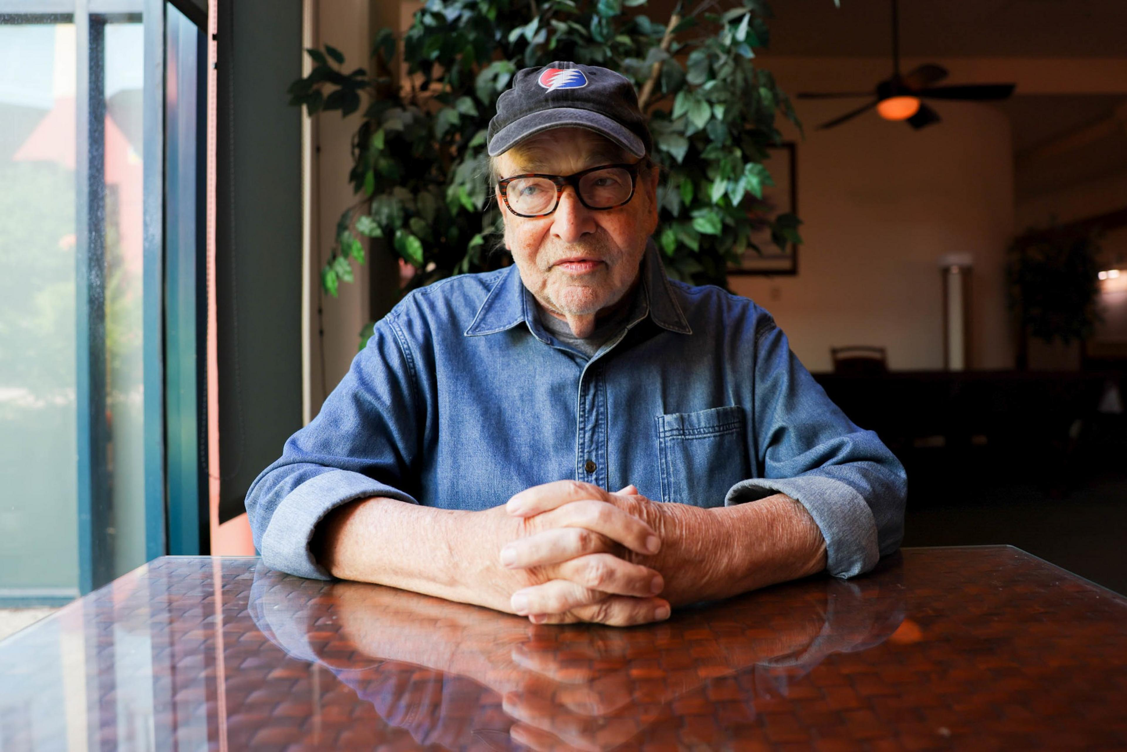 An older man wearing glasses, a blue denim shirt, and a black cap sits at a wooden table with his hands clasped.