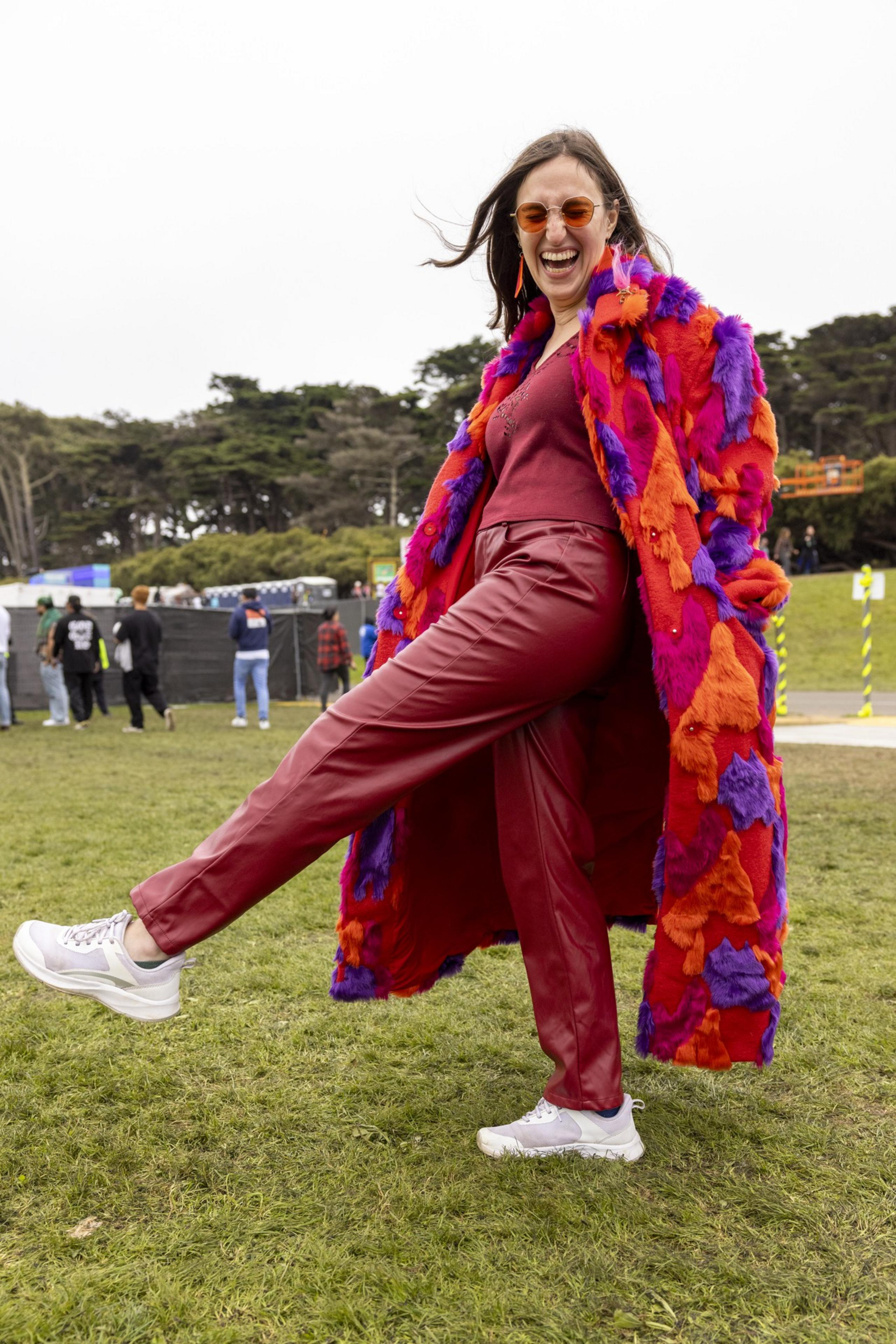 A joyful woman stands on grass, kicking up a leg playfully. She wears orange sunglasses, a vibrant furry coat, red pants, and white sneakers, with trees in the background.