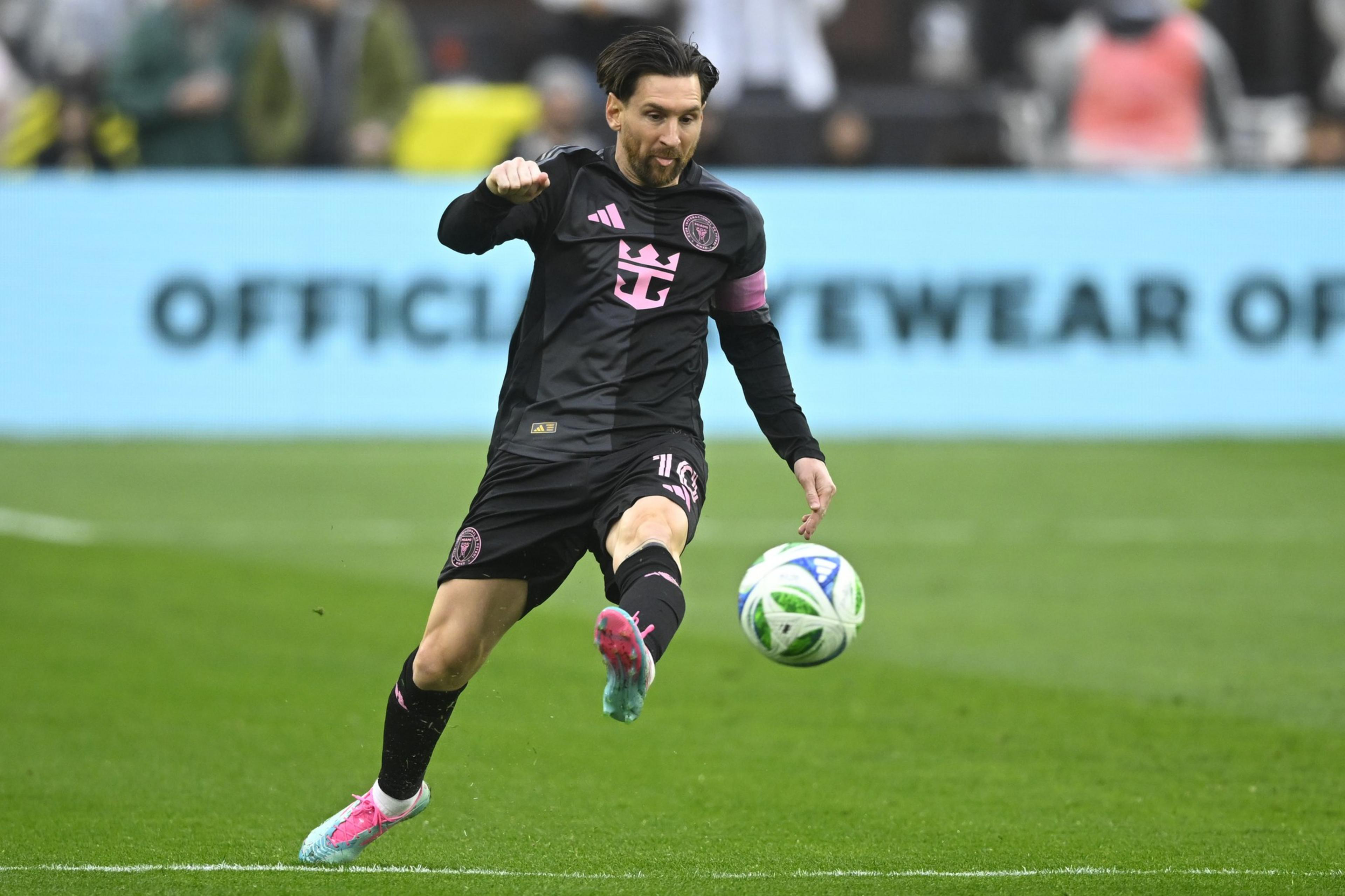 A soccer player in a black and pink uniform kicks a ball on a grassy field. The background is a blurred advertisement and spectators.