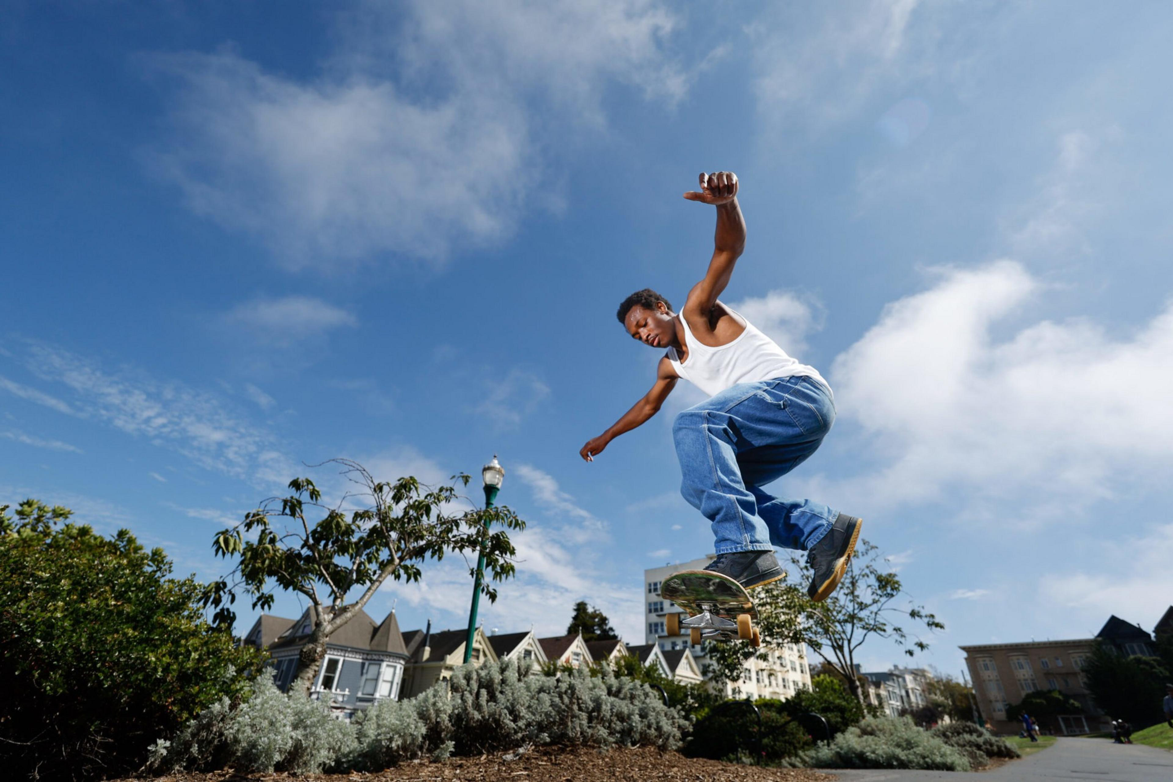 A man wearing a white tank top and blue jeans is mid-air performing a skateboard trick in an urban park under a blue sky.