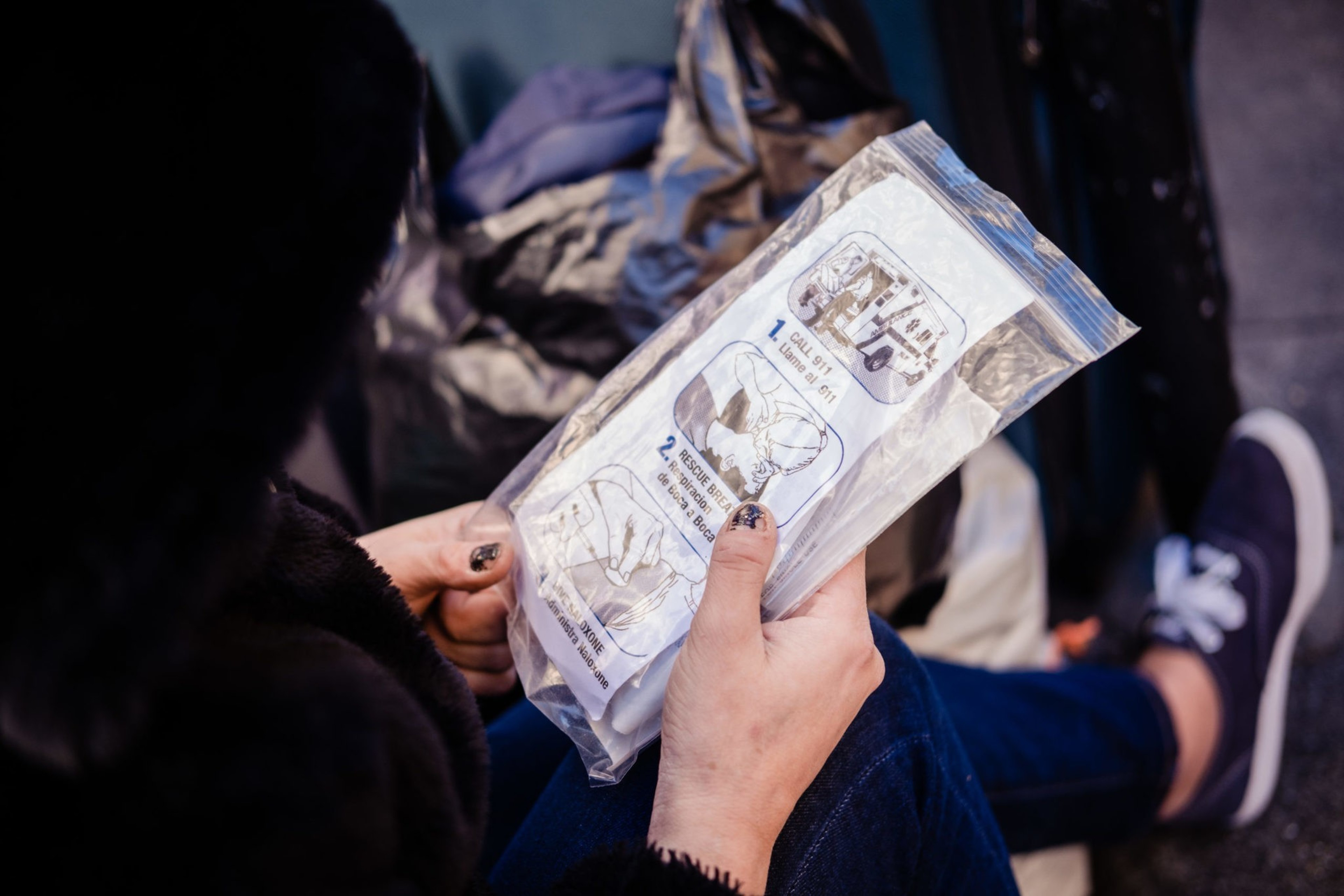 A person is holding a sealed plastic bag containing a medical kit with illustrated instructions. Their nails are painted black, and casual shoes are visible.