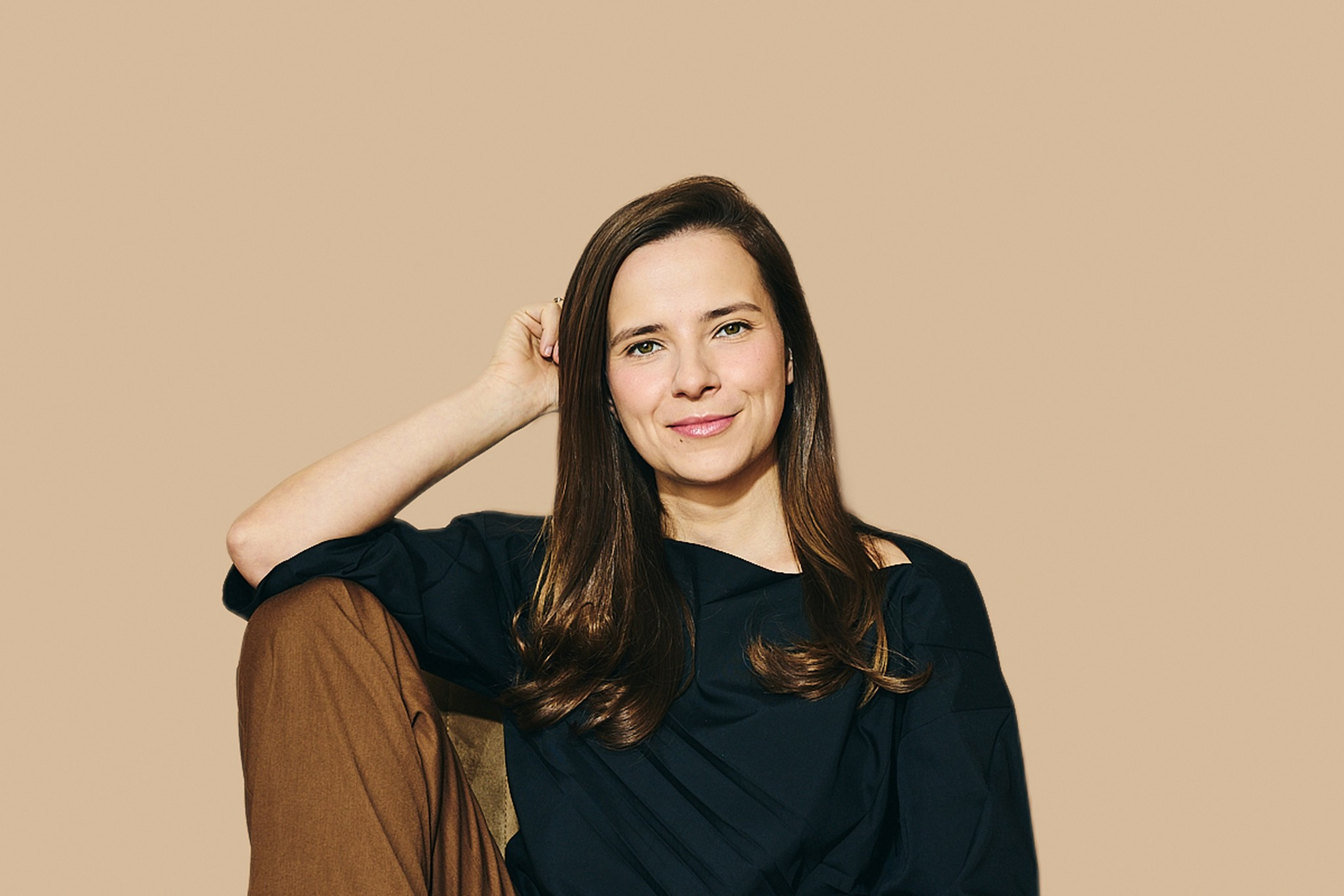 A woman with long brown hair wears a black top and brown pants, smiling ...