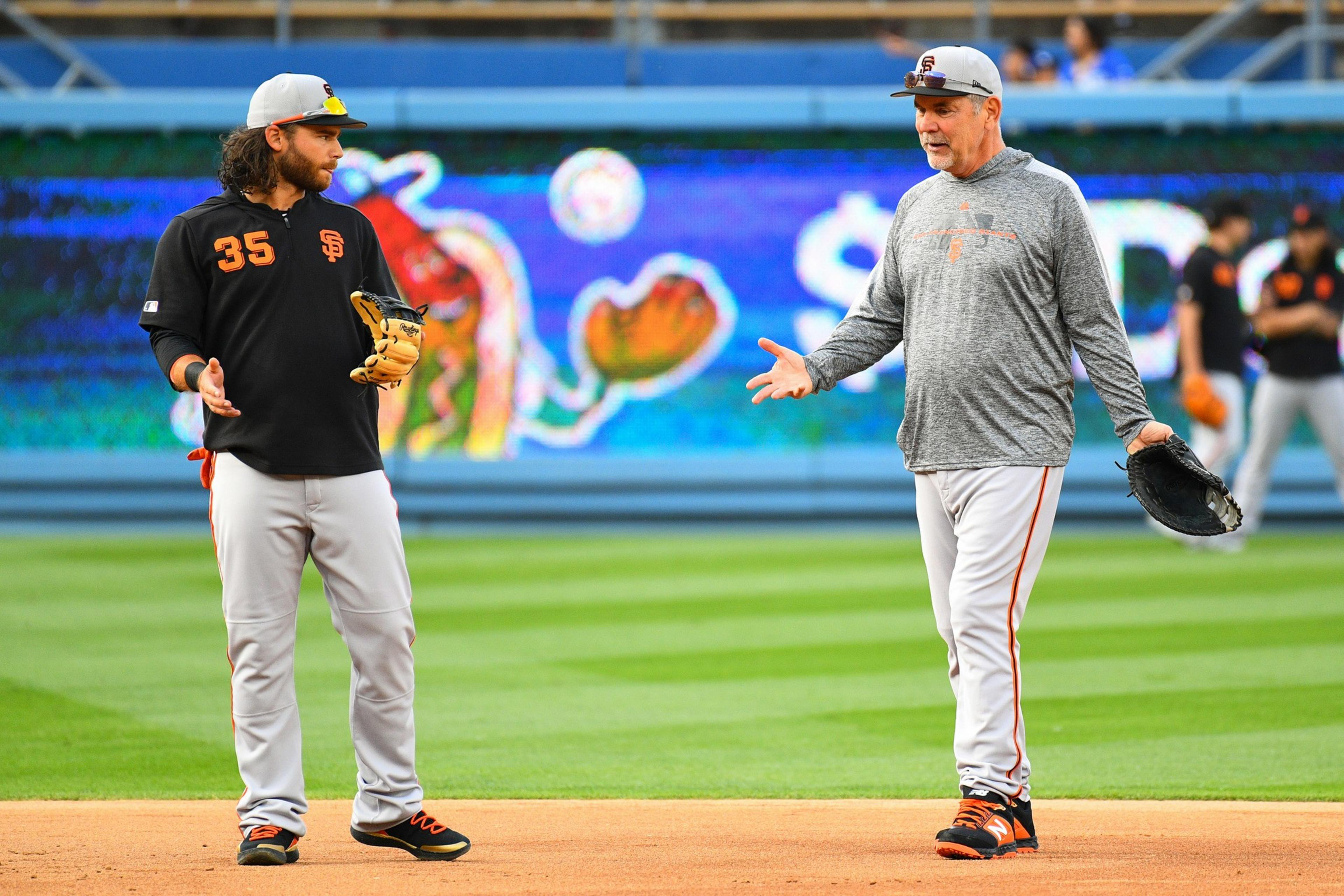 Two baseball players stand on a field, wearing uniforms with orange accents. One holds a glove and gestures, while the other appears to be listening.