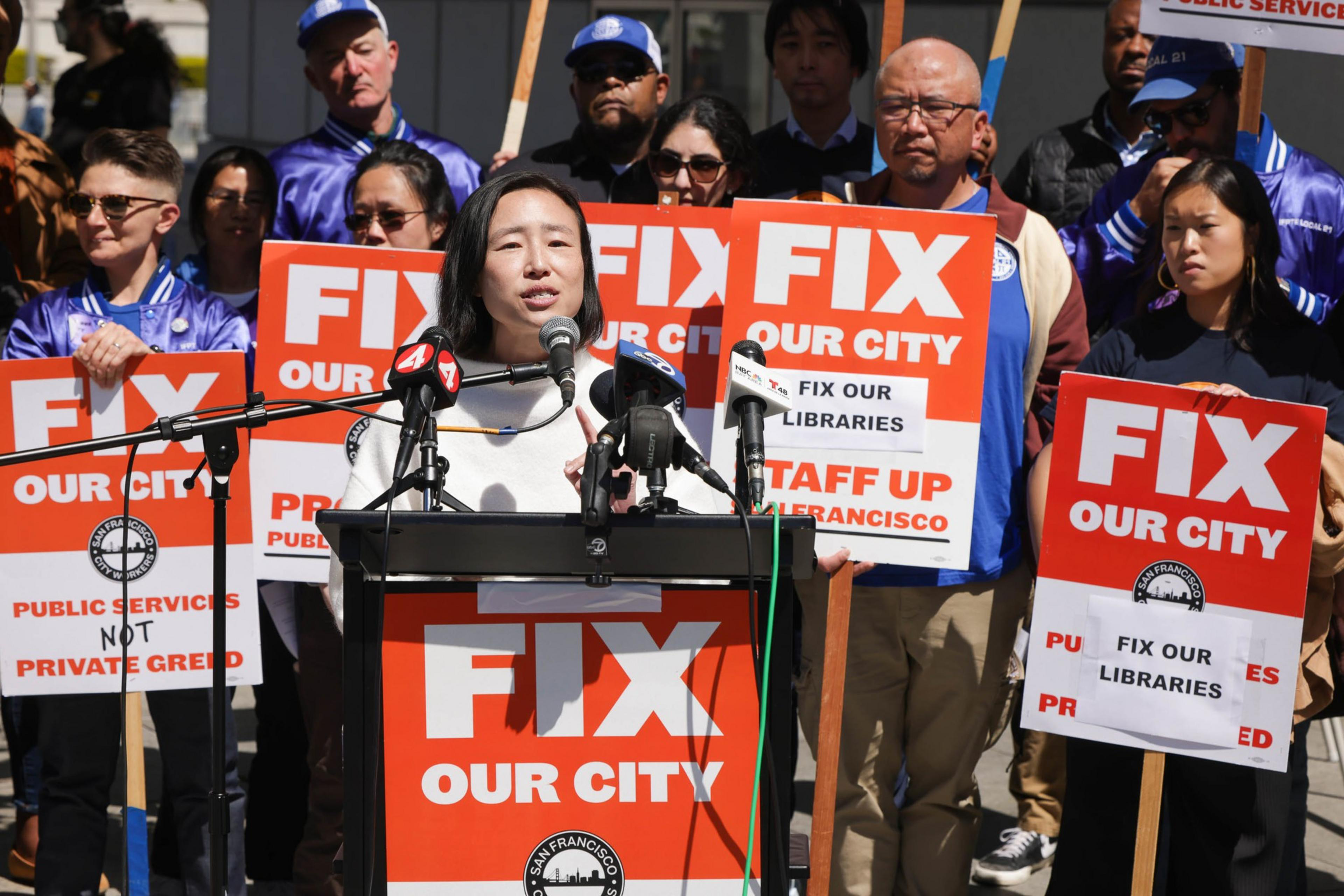 A woman speaks at a podium surrounded by people holding orange signs that say "FIX OUR CITY" and call to fix libraries and public services.