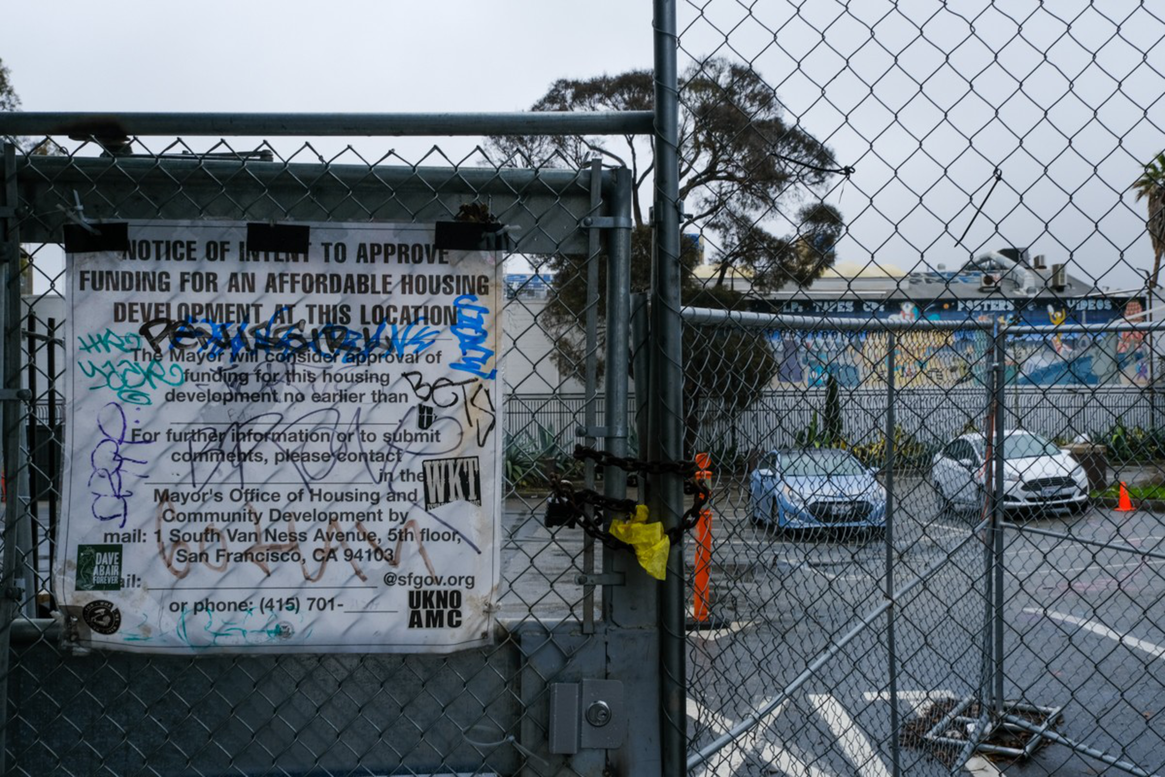 A chain-linked fence with a locked gate and a partly graffiti-covered notice about affordable housing funding in front of an empty parking lot and mural walls.