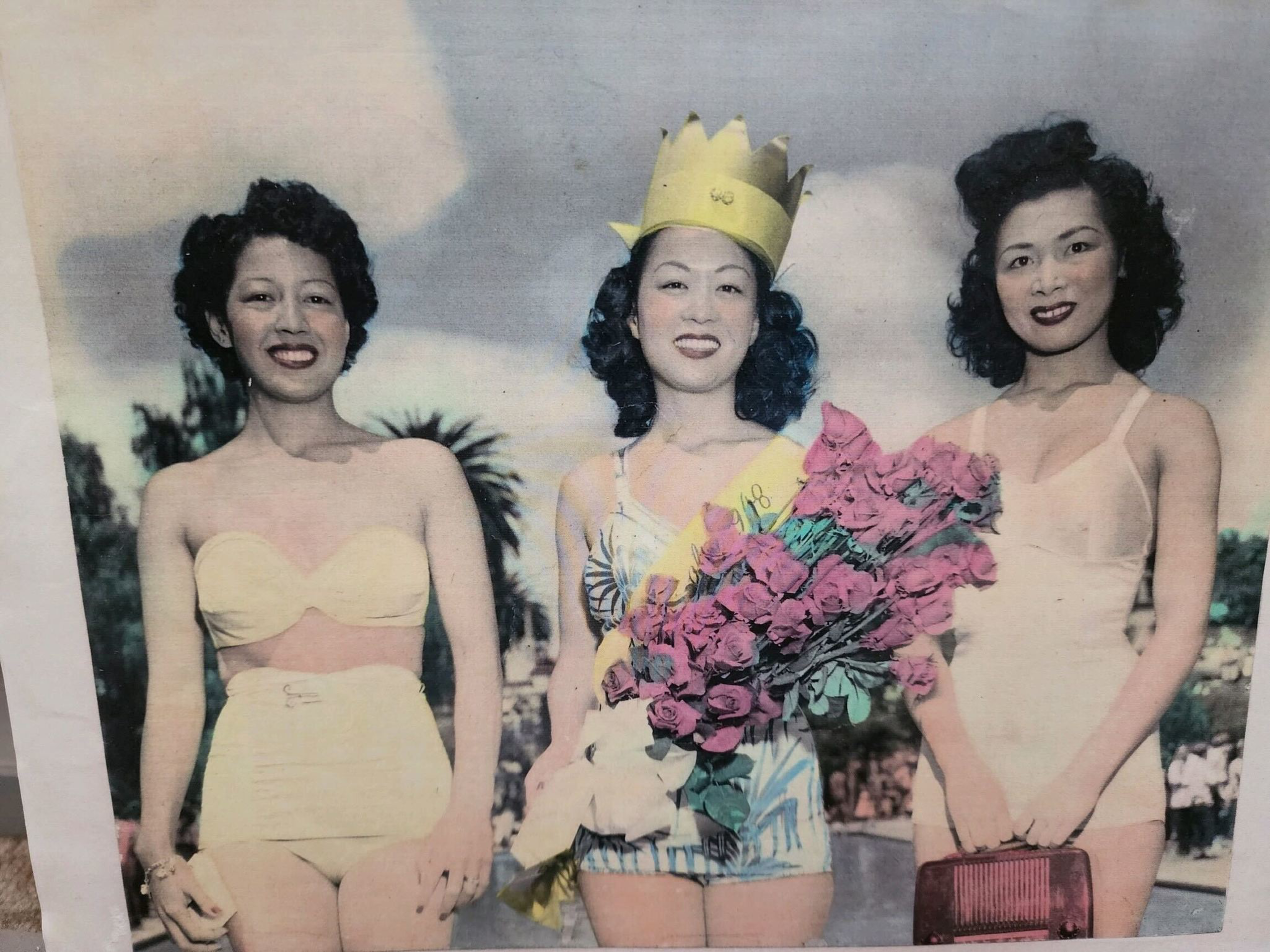 Three women in vintage swimsuits pose outdoors, the center woman wears a paper crown and sash, holding a bouquet of pink roses.