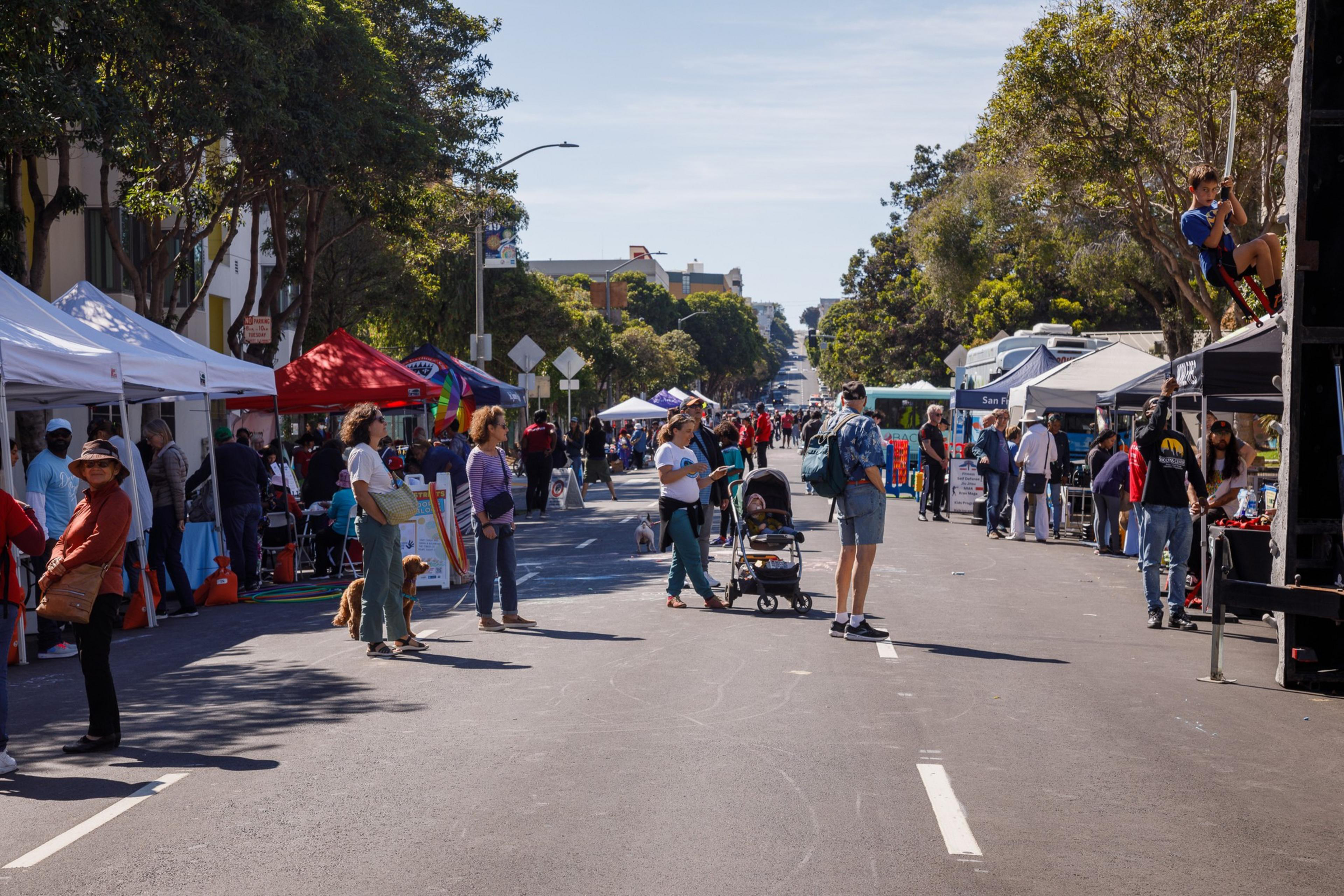 A bustling outdoor street market with tents, visitors, and a child climbing a structure on a sunny day.