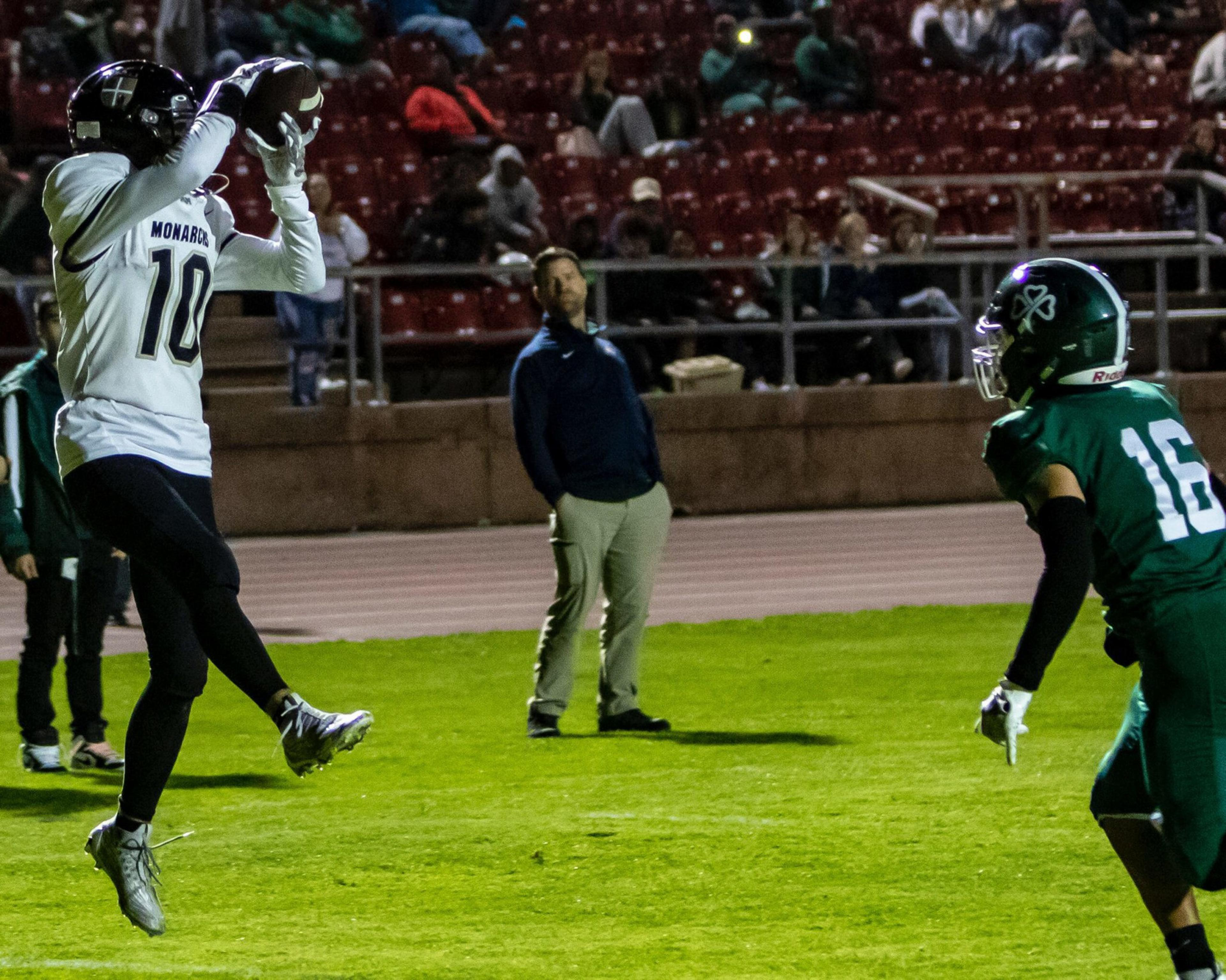 A football player in a white jersey leaps to catch the ball as a player in a green jersey approaches on a grassy field during a night game.