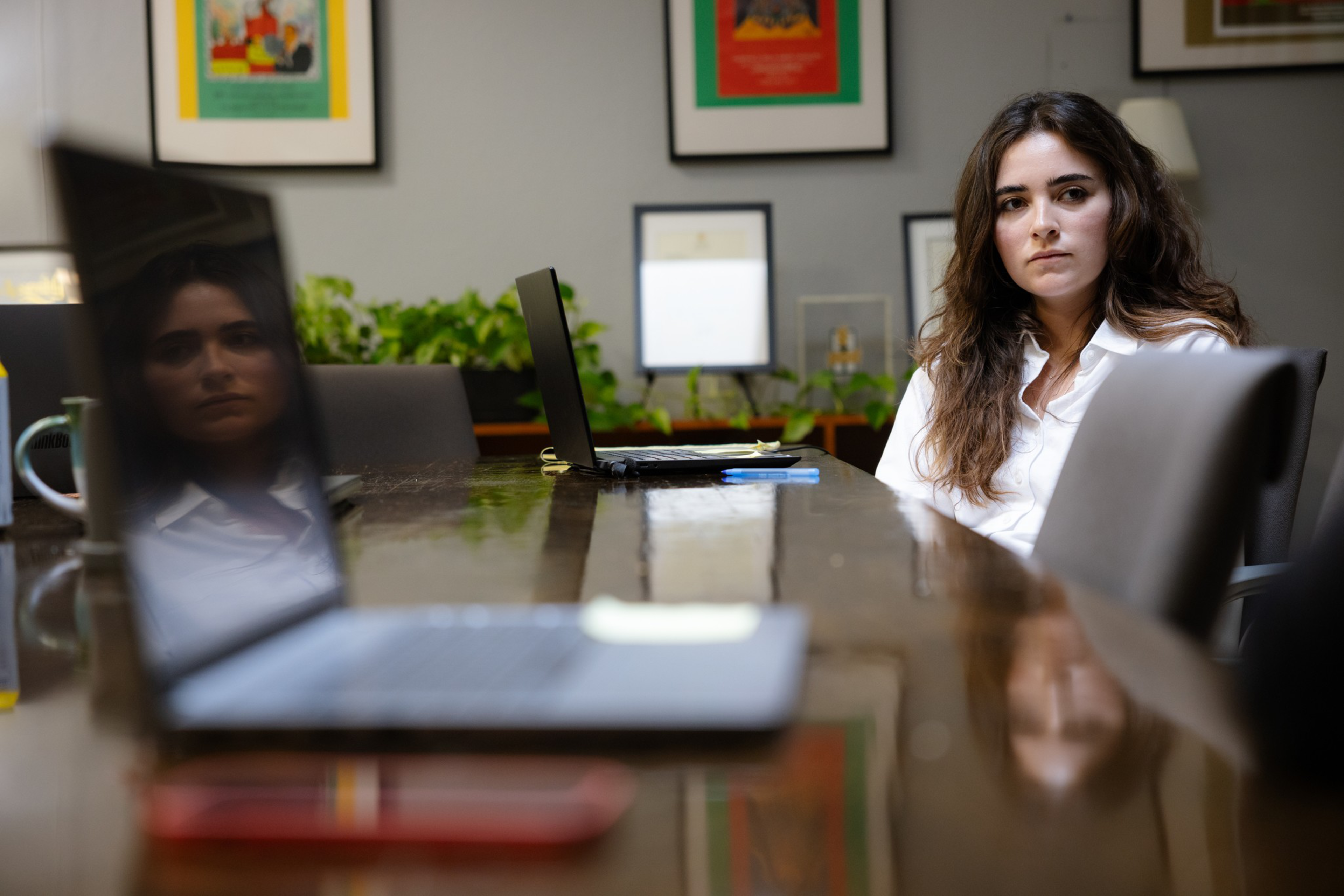 A woman with long brown hair and a white shirt sits at a conference table, looking pensively toward an open laptop reflecting her face.