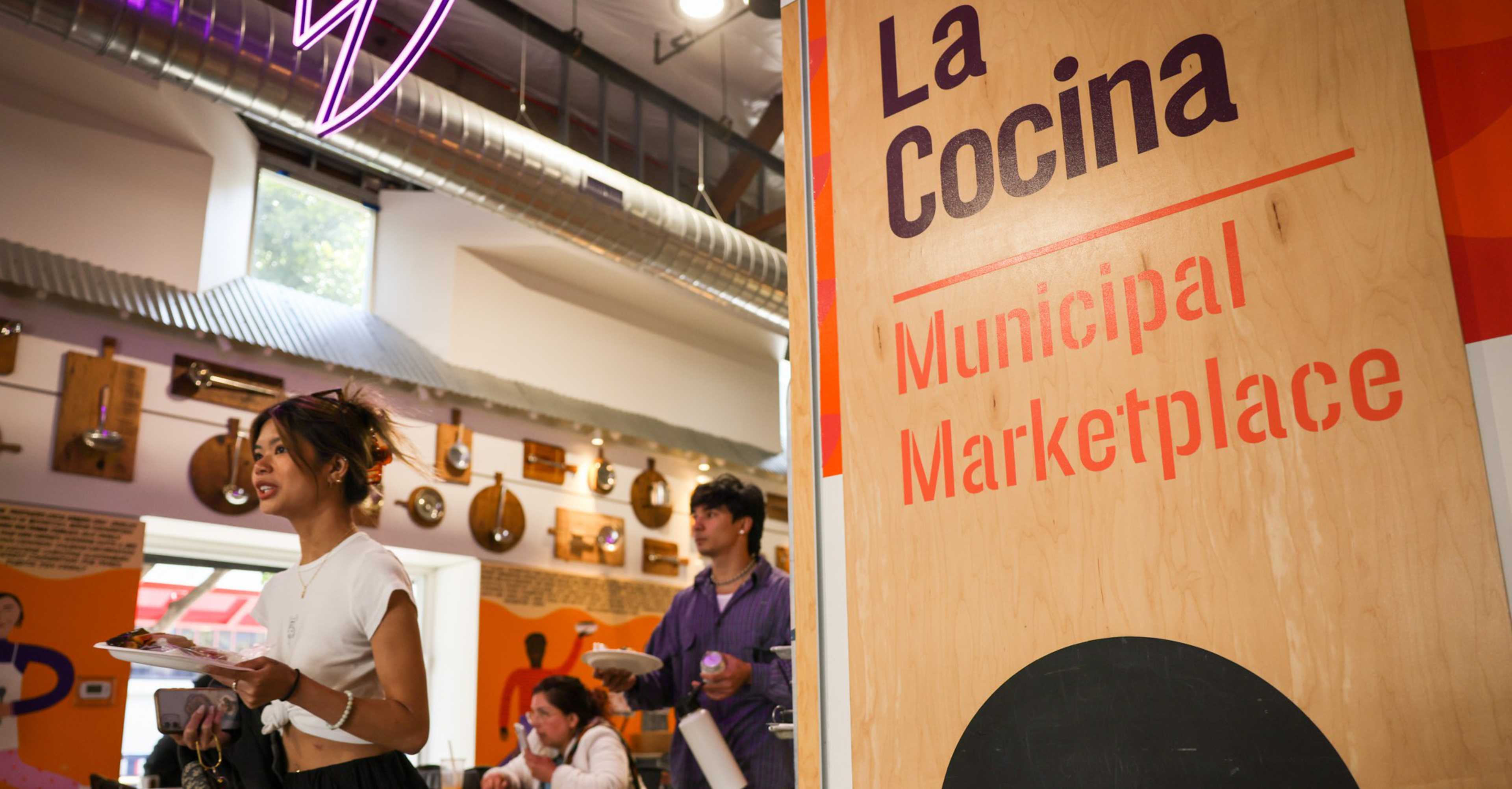 People carrying plates walk and sit inside a marketplace area with guitars hanging on the wall, next to a wooden sign that reads "La Cocina Municipal Marketplace."