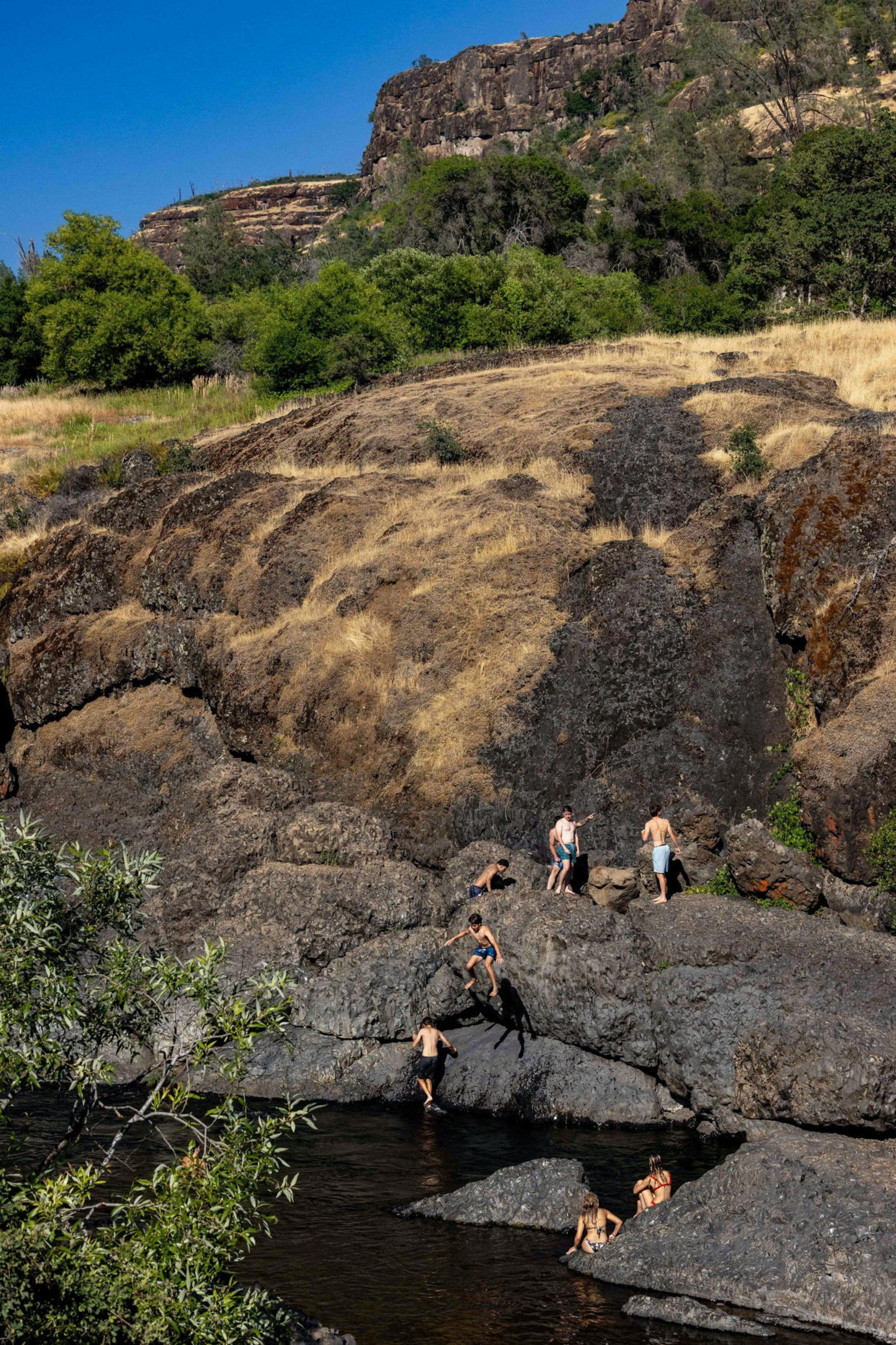 People are climbing rocky terrain near a river with a backdrop of hills, trees, and a clear blue sky.