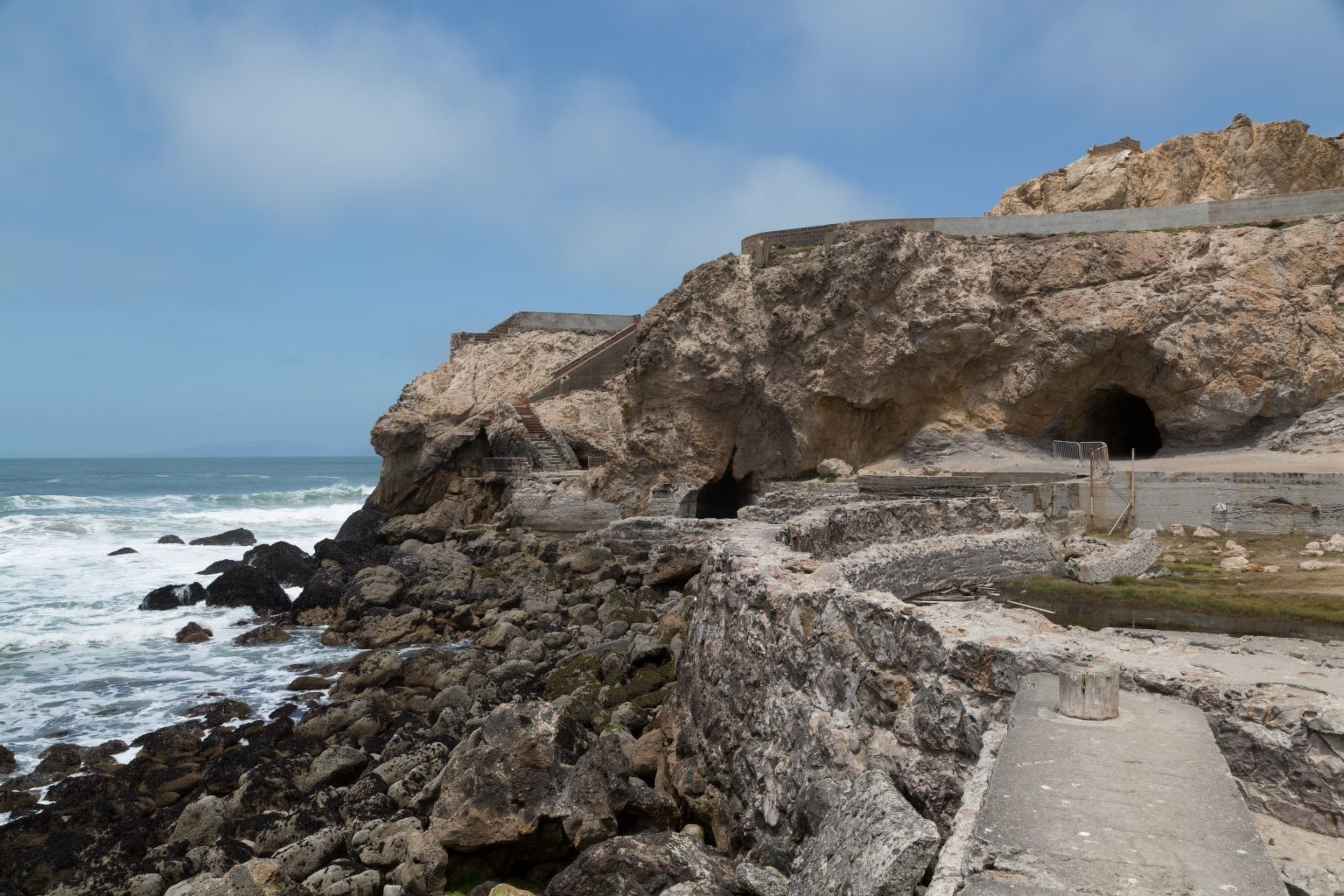 The image shows a coastal scene with rocky cliffs, sea caves, and ocean waves crashing against the rocks under a partly cloudy sky. A path and stairs are visible on the cliff.