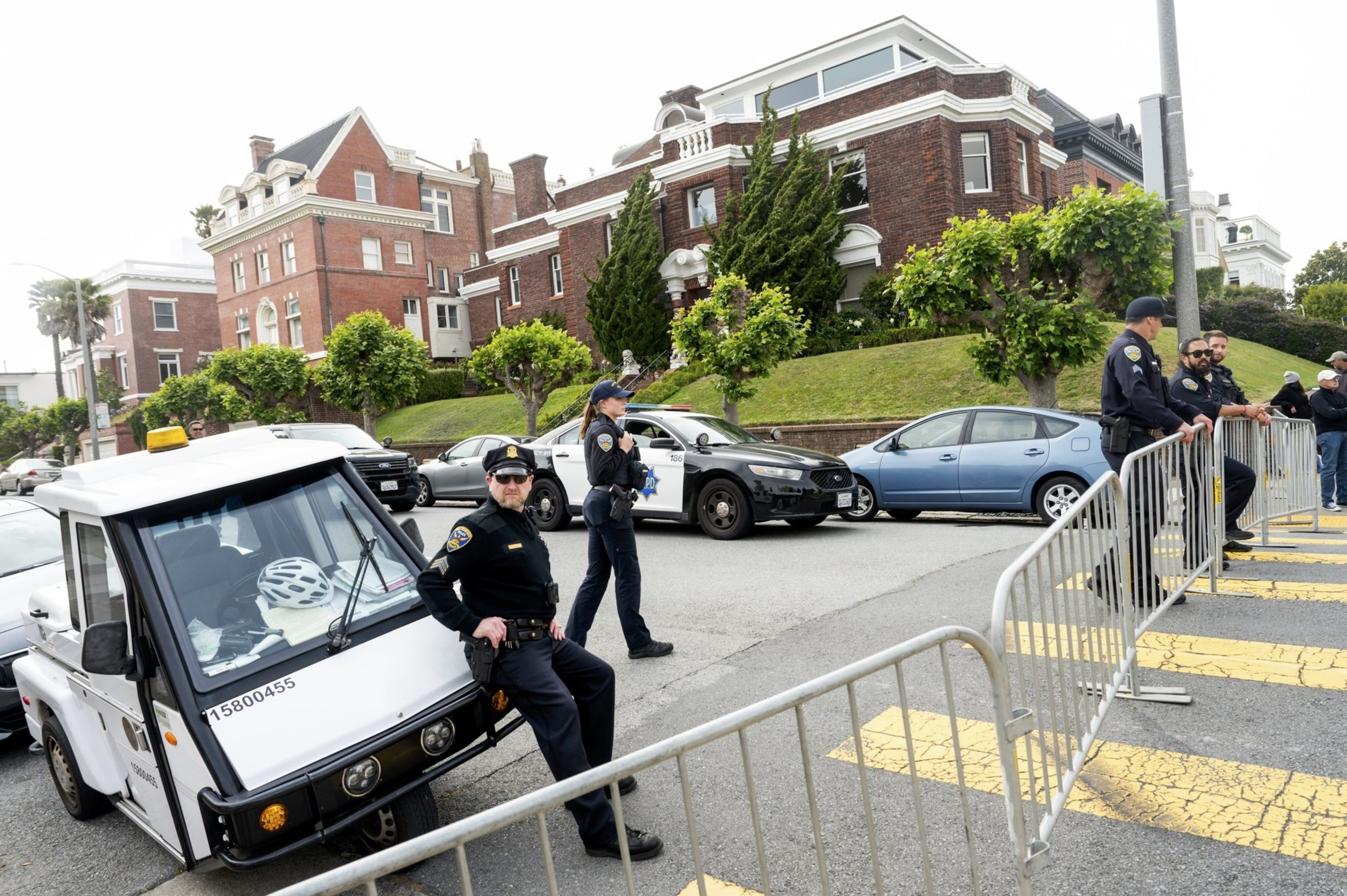 Police officers stand by barricades on a residential street lined with large brick houses, overseeing parked cars, including a white utility vehicle and patrol car.