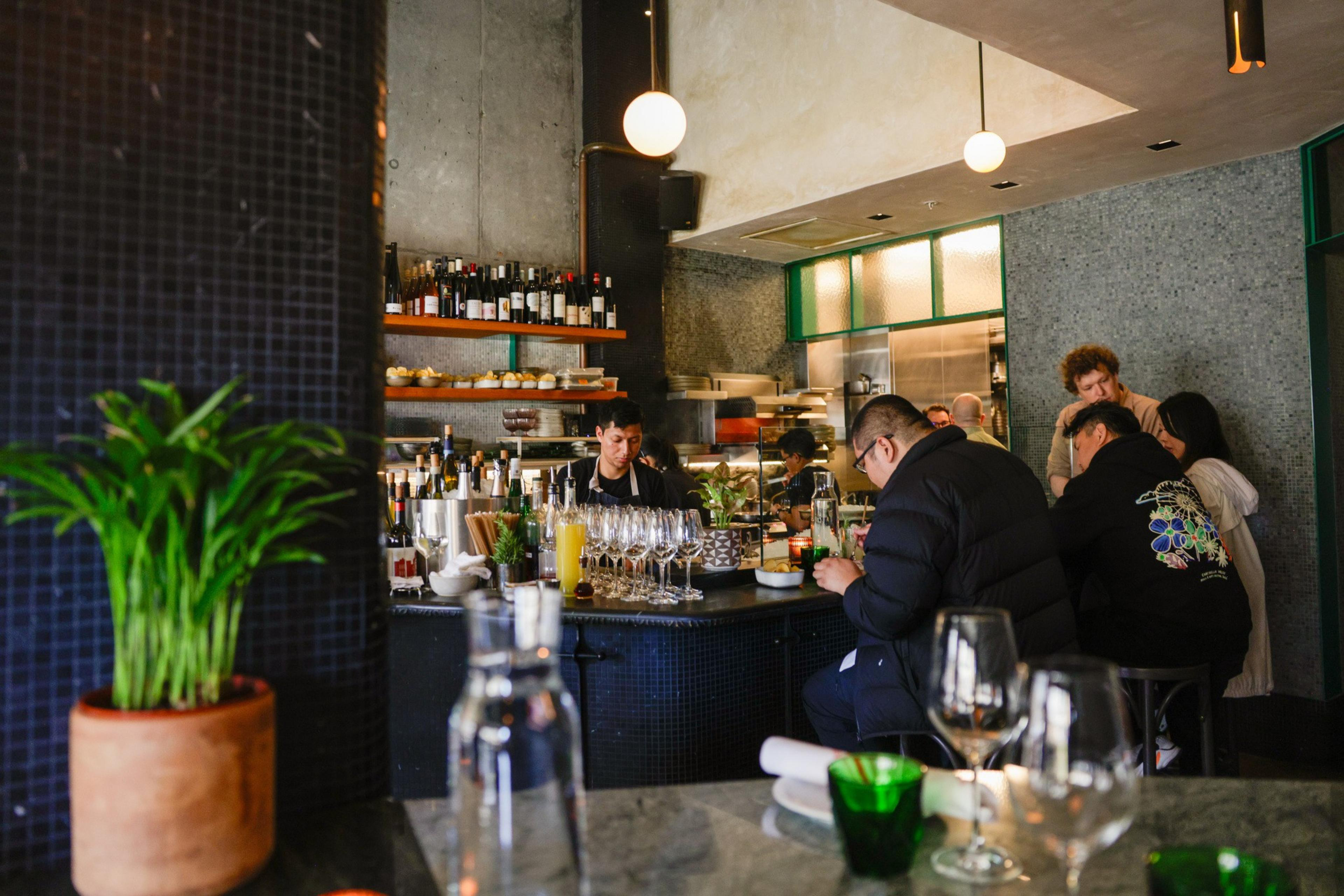 A cozy bar scene with people seated at the counter. There are shelves with wine bottles, glasses set out, and a potted plant on the side.