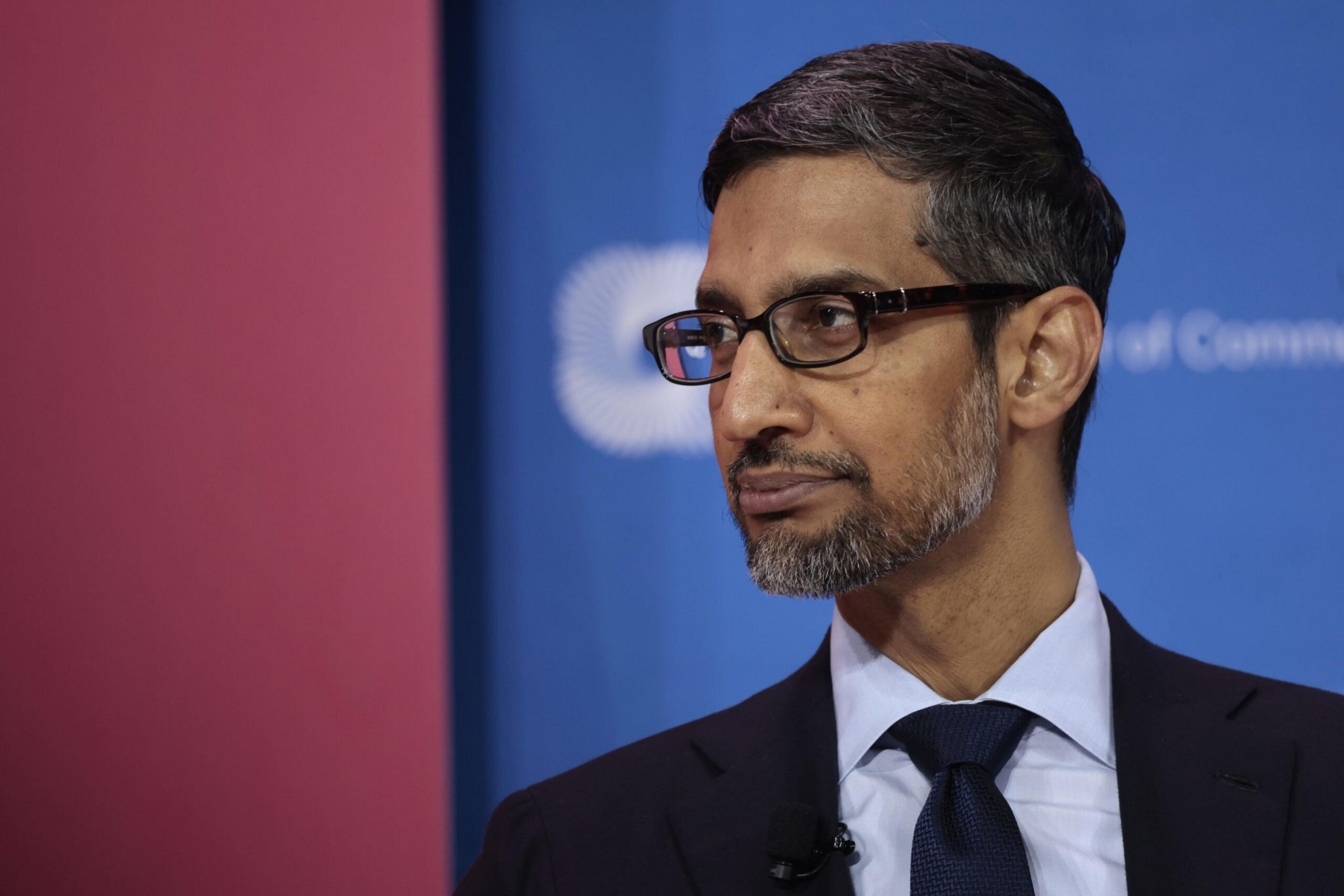 A man with short dark hair and a salt-and-pepper beard wears glasses, a navy suit, light blue shirt, and dark tie, looking thoughtfully to the side.