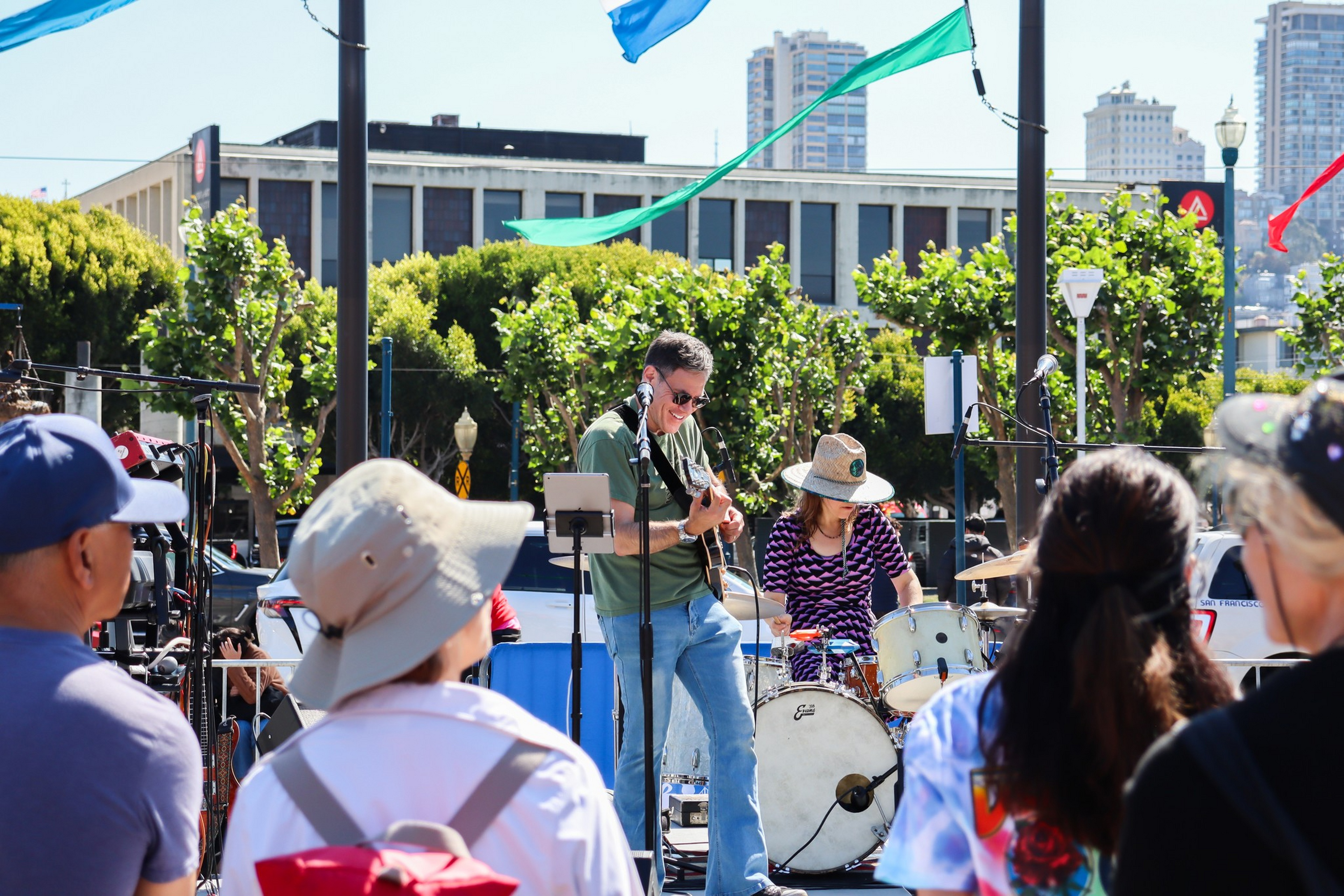 A man plays guitar and a woman plays drums outdoors, performing for an attentive crowd on a sunny day with colorful flags and city buildings in the background.