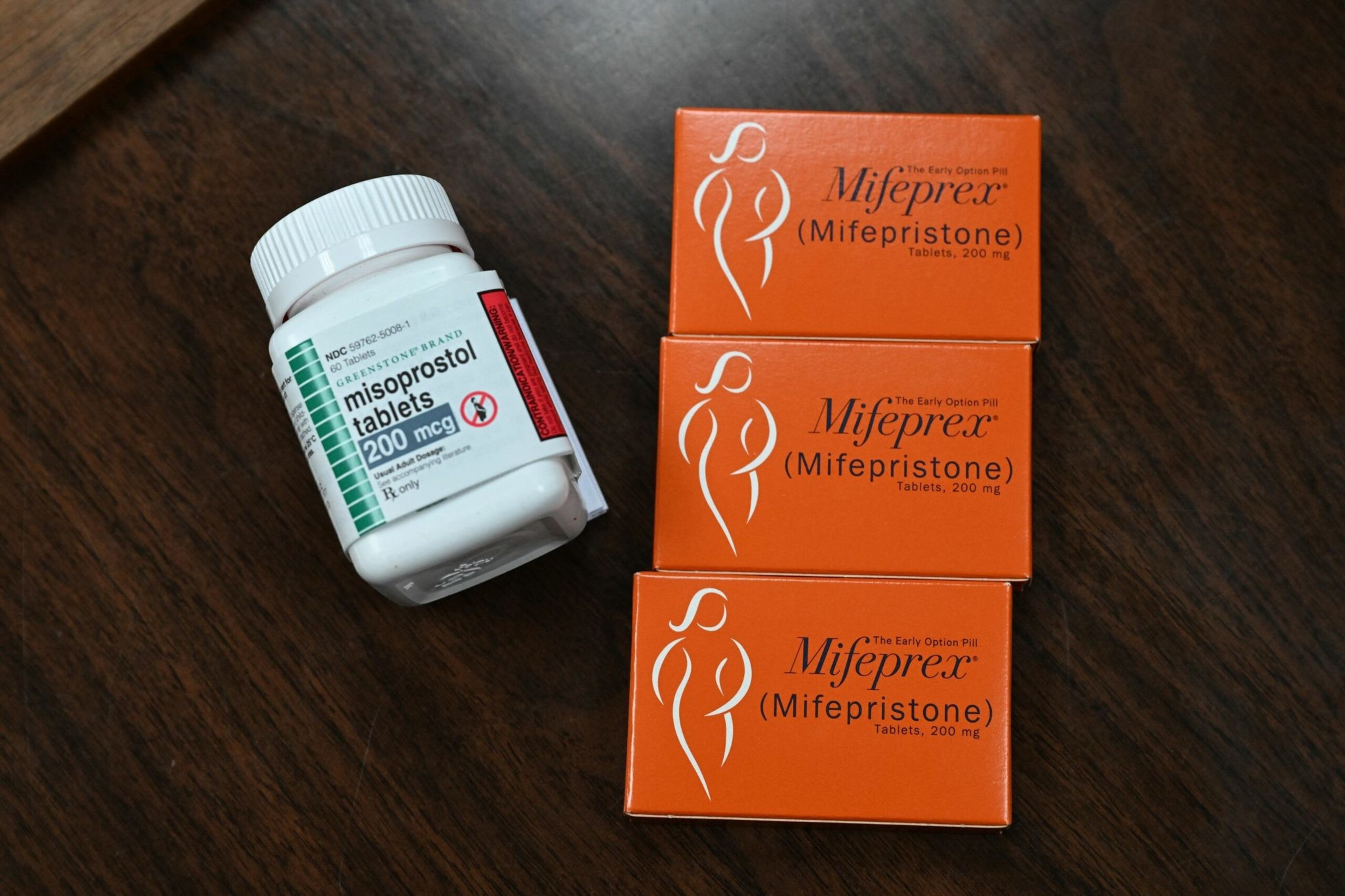 A white bottle labeled Misoprostol tablets 200 mcg stands beside three orange boxes labeled Mifeprex (Mifepristone) tablets 200 mg on a wooden surface.