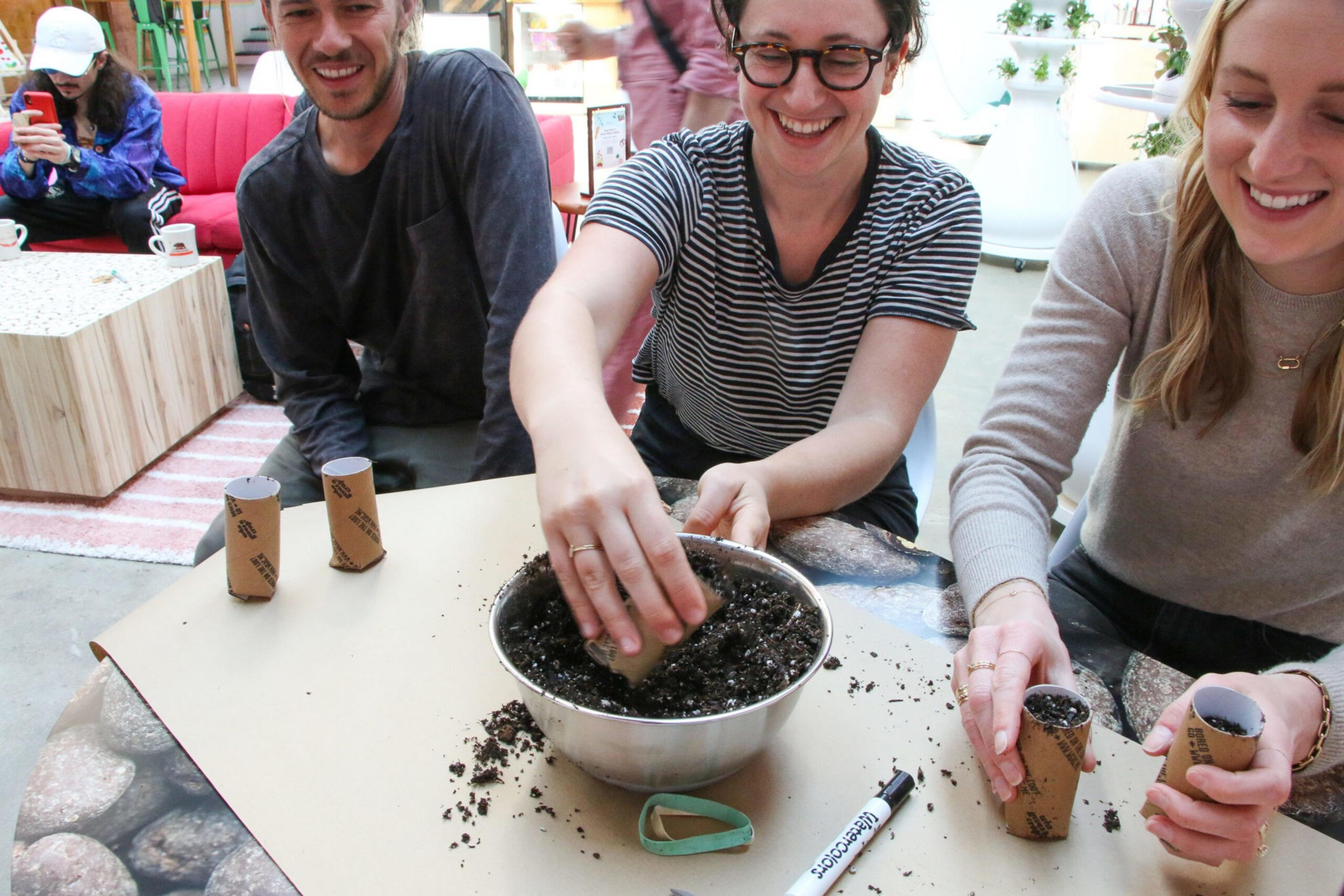 Three people are smiling and filling small cardboard tubes with soil from a metal bowl on a table, preparing for planting.