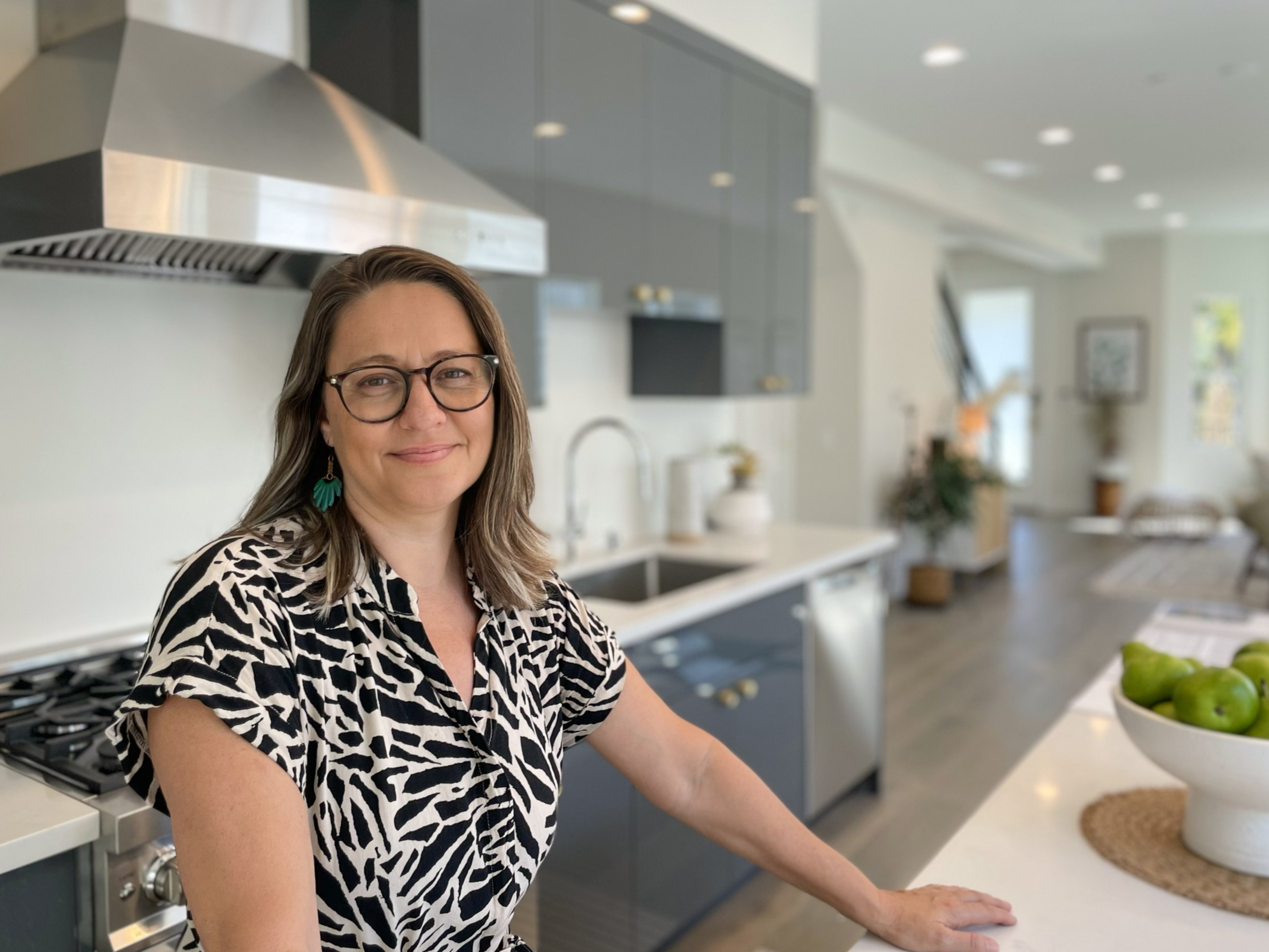 A woman with glasses and patterned shirt stands in a modern kitchen, smiling. The kitchen features stainless steel, dark cabinets, and a white countertop with green apples.