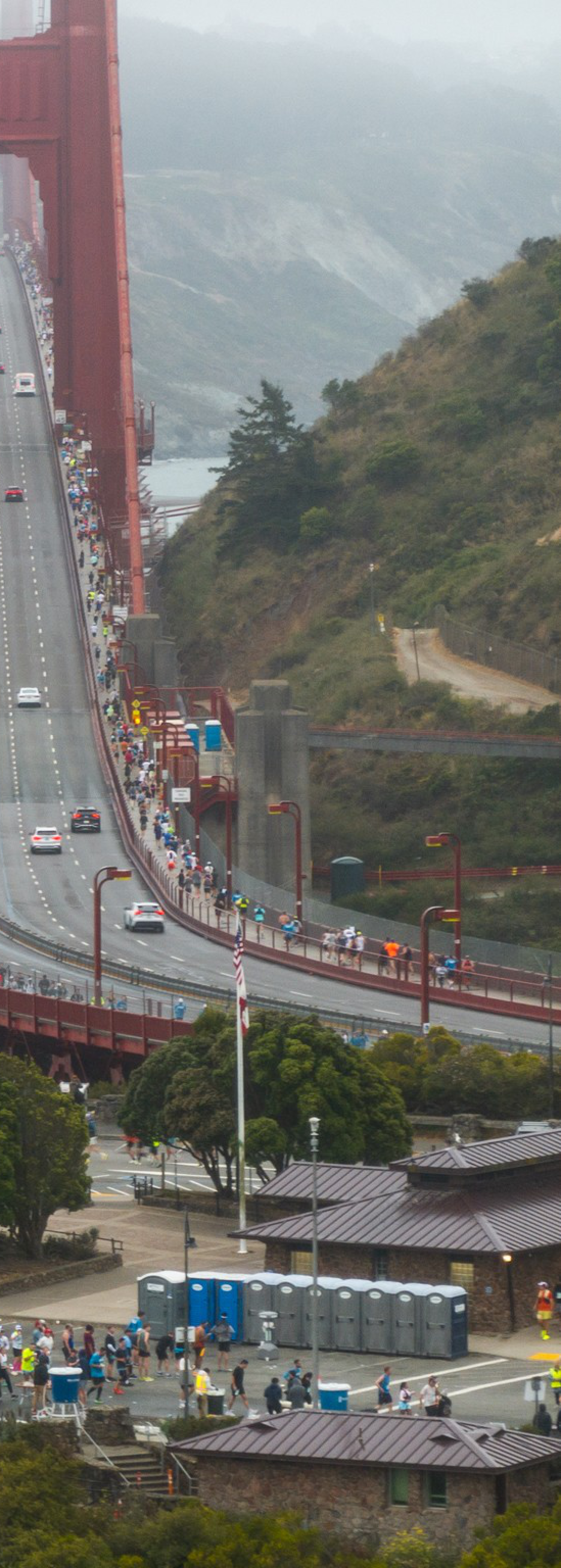 A large group of people is gathered at one end of the Golden Gate Bridge, with some cars driving across it. The scene overlooks water and a foggy, tree-filled background.