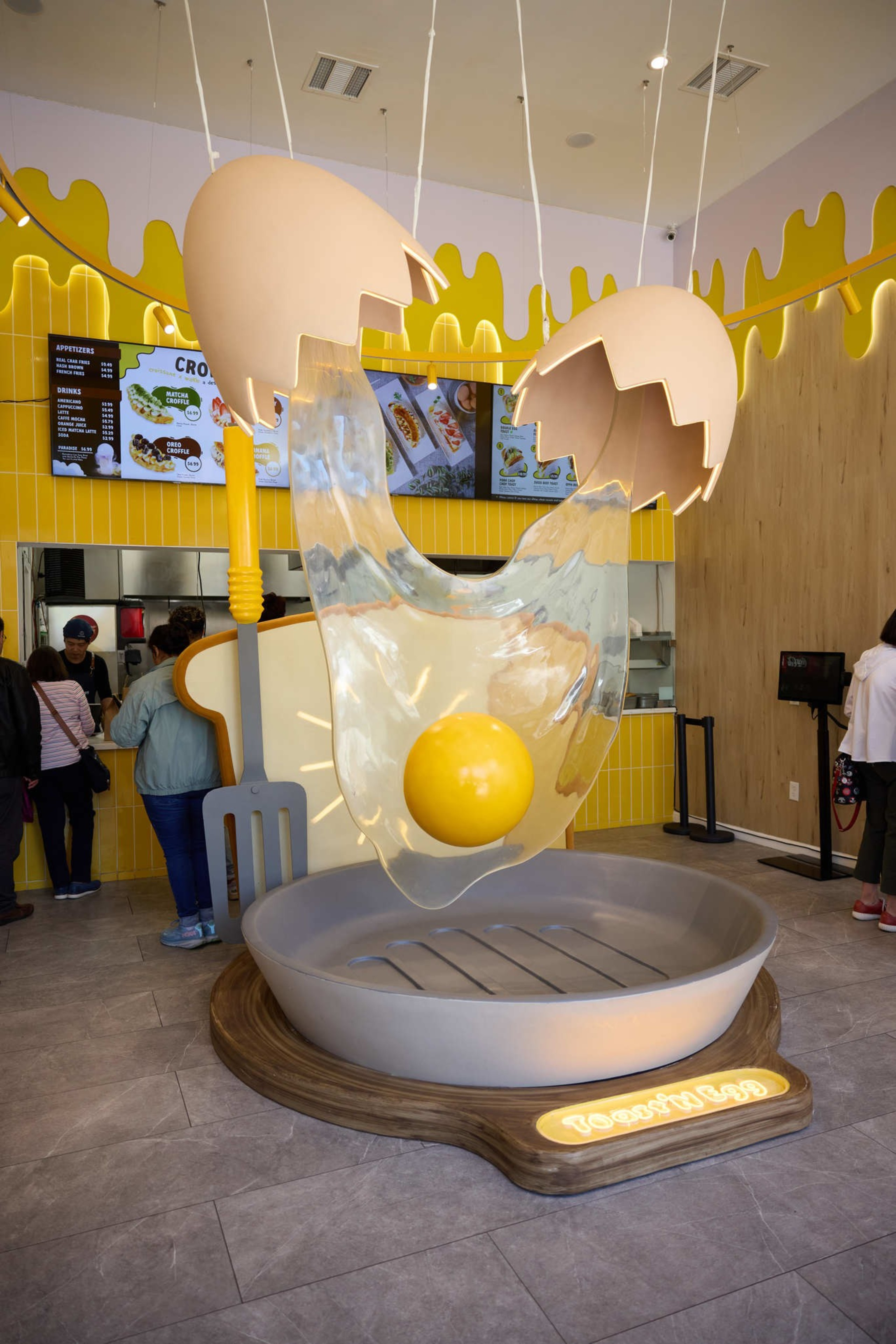 The image shows a restaurant with a giant egg sculpture hanging from the ceiling, with a yolk falling into a large skillet. Customers are lined up by the counter.