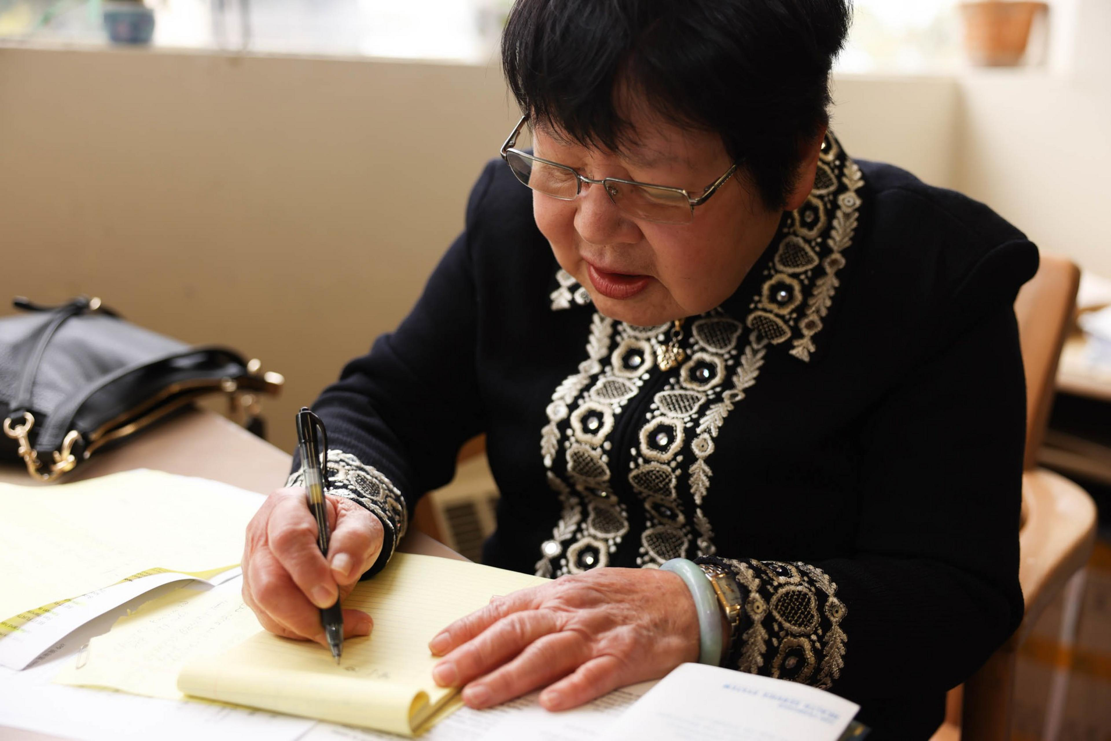 An elderly woman wearing glasses and a black outfit with intricate white patterns writes on a yellow notepad at a table, with a black purse nearby.