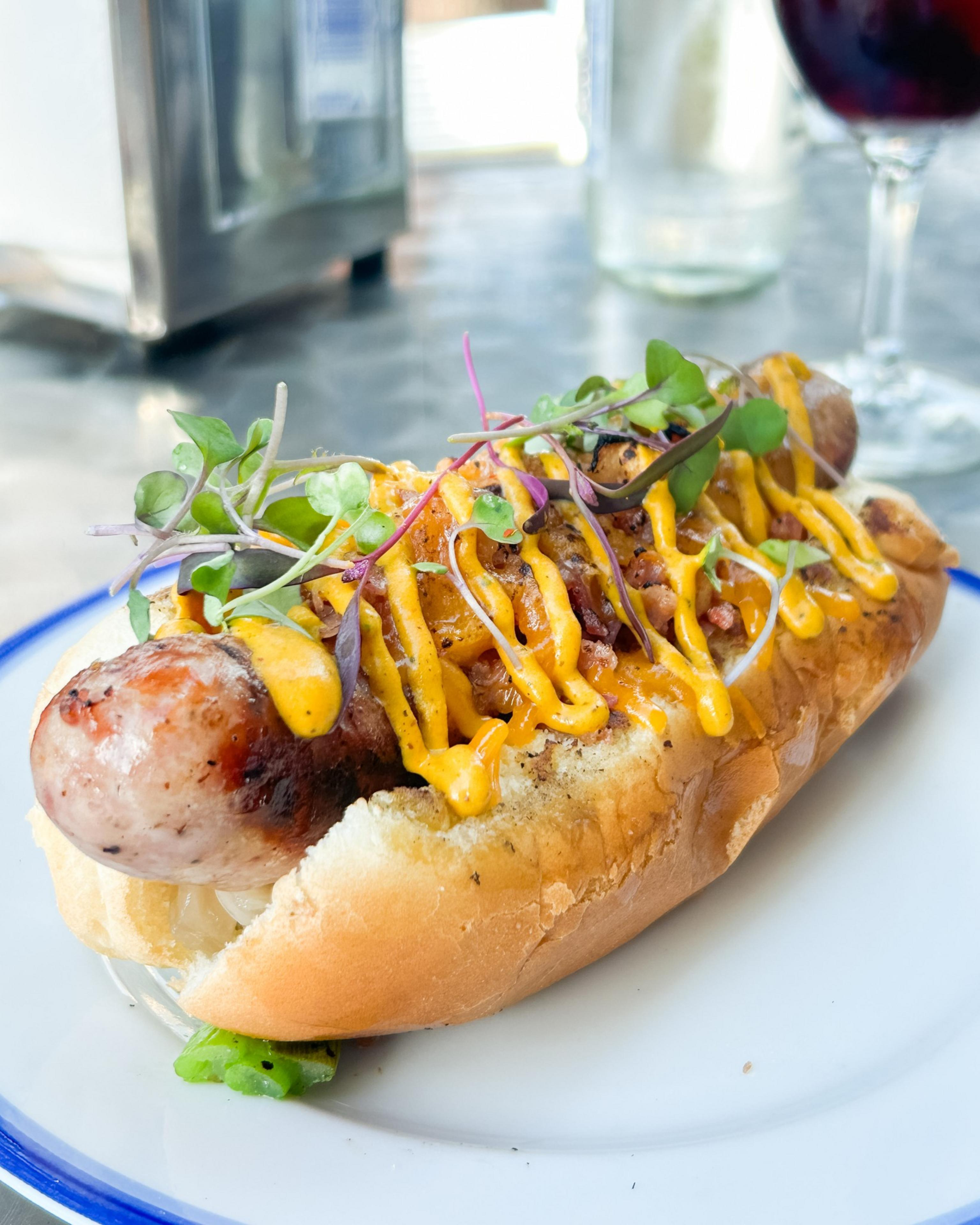 A sausage in a bun is topped with mustard, bacon bits, and fresh greens. It's served on a white plate with a blue rim, next to a glass and metal container.