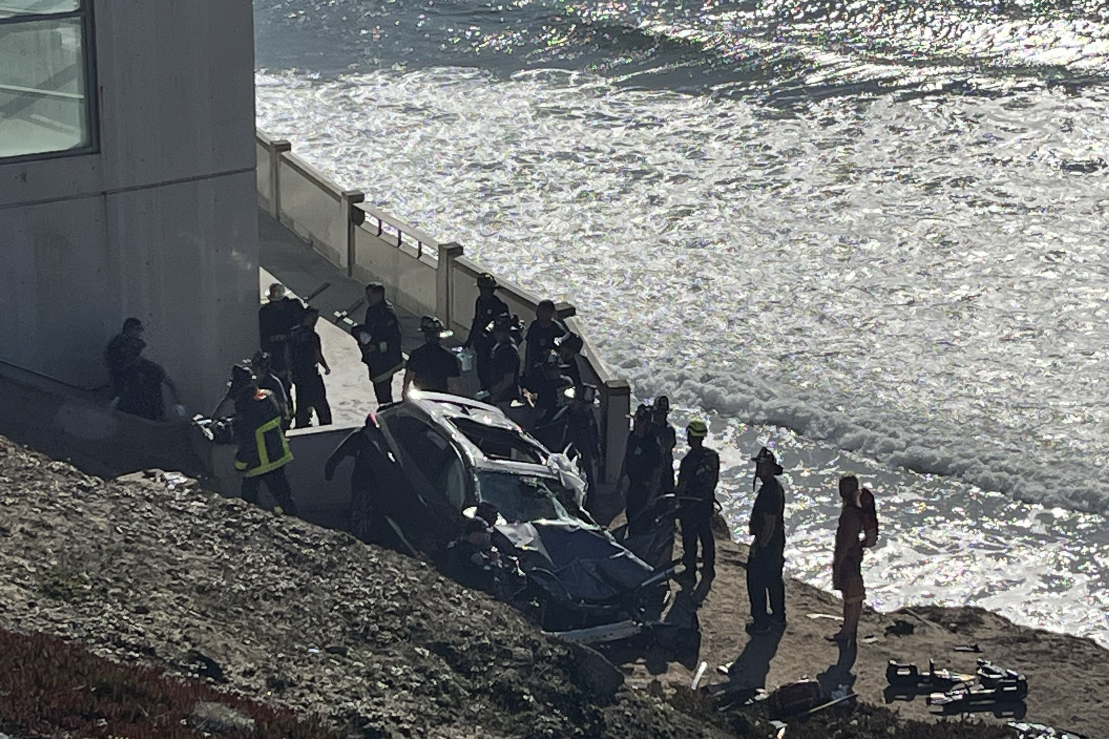 The image shows a car crash scene by the seaside, with emergency personnel gathered around a badly damaged vehicle at the base of a concrete structure.
