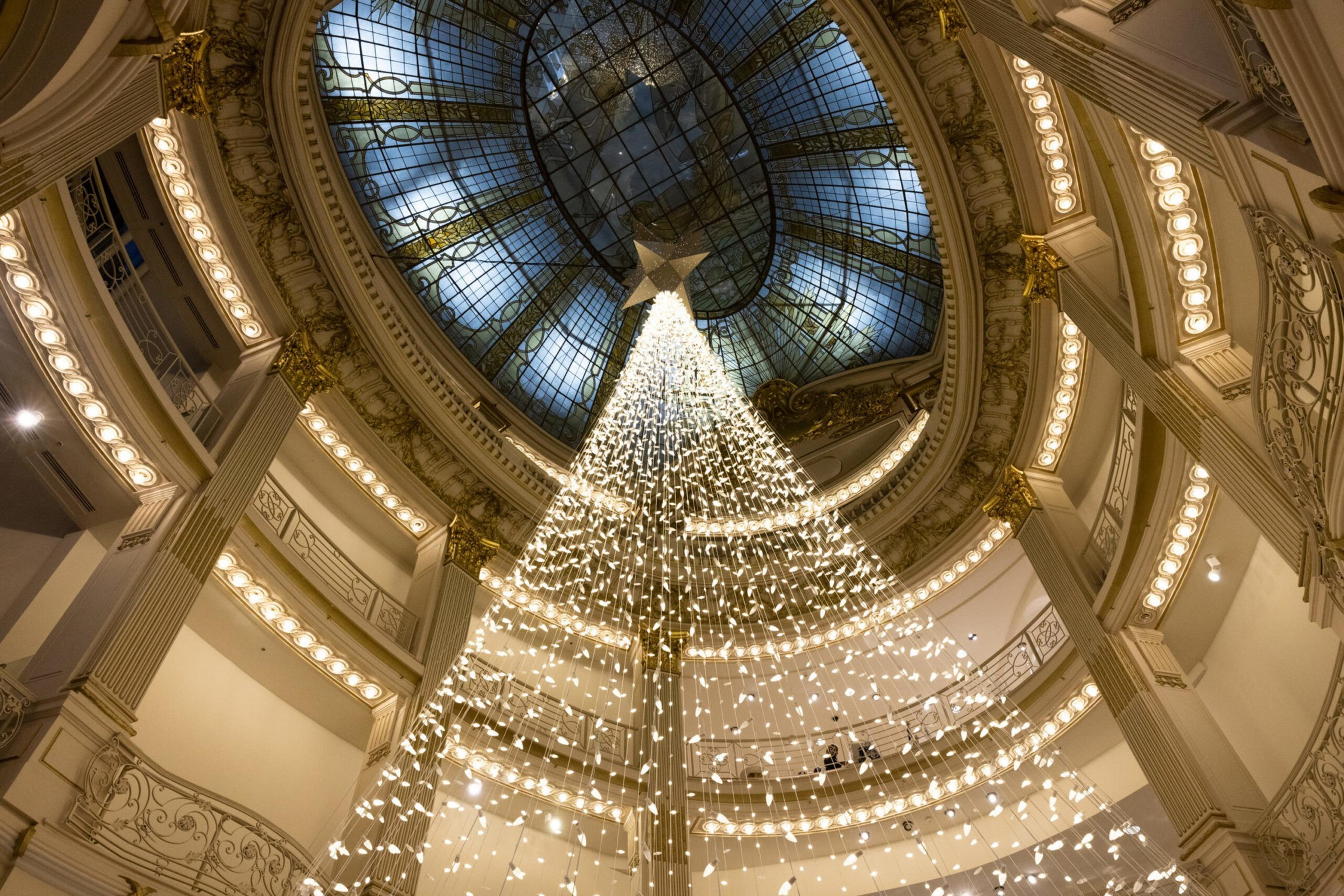 A large cone-shaped Christmas tree made of cascading white lights hangs under an ornate glass dome inside a grand, multi-level building with intricate railings.