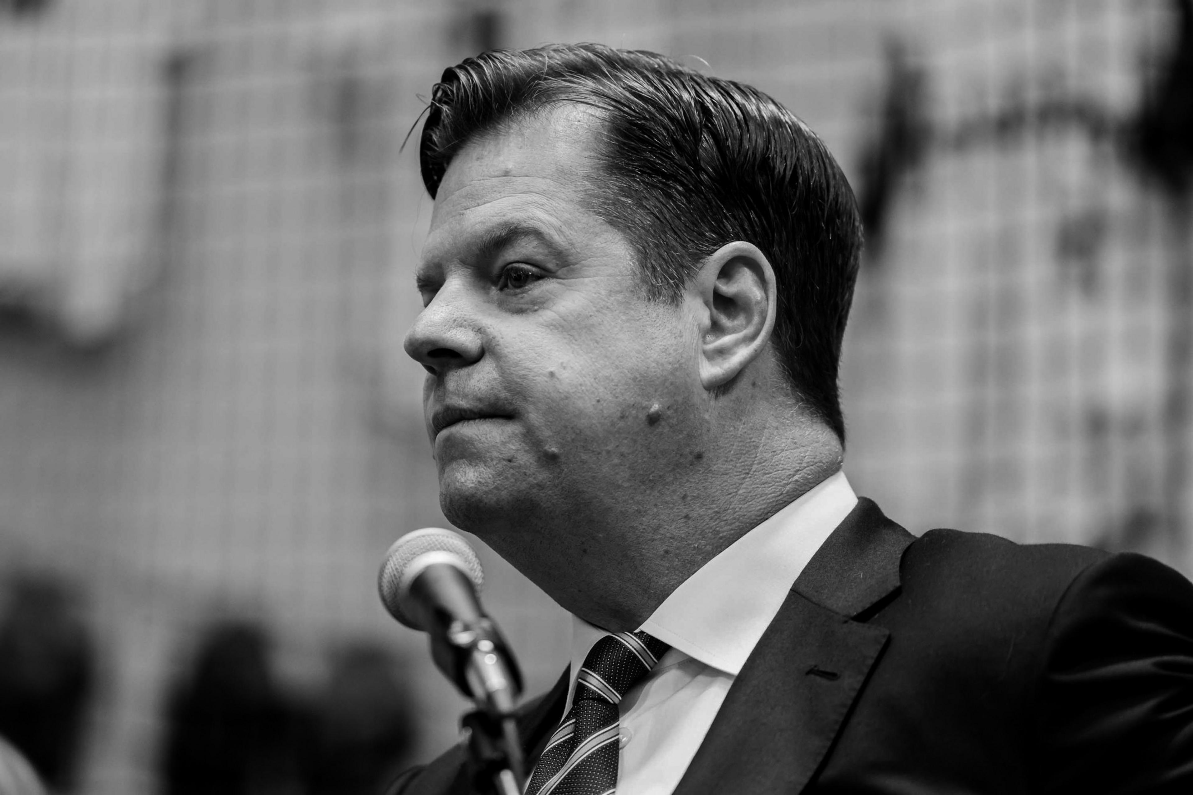A black and white photo of a man in a suit, with a microphone in the foreground. He has slicked-back hair and a serious expression.