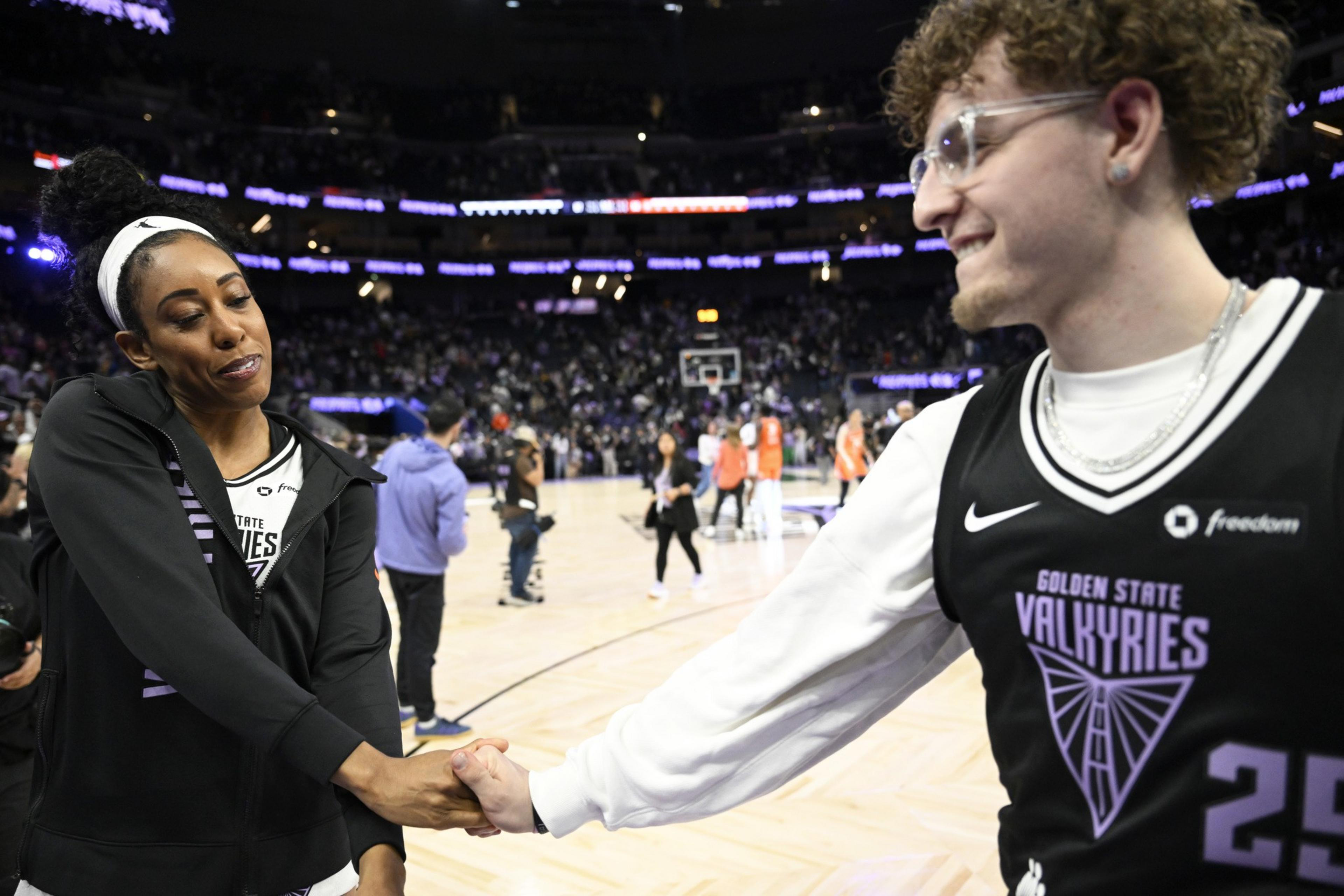 Two people are on a basketball court shaking hands, with a crowd in the background. One wears a &quot;Golden State Valkyries&quot; jersey and the other a black jacket.