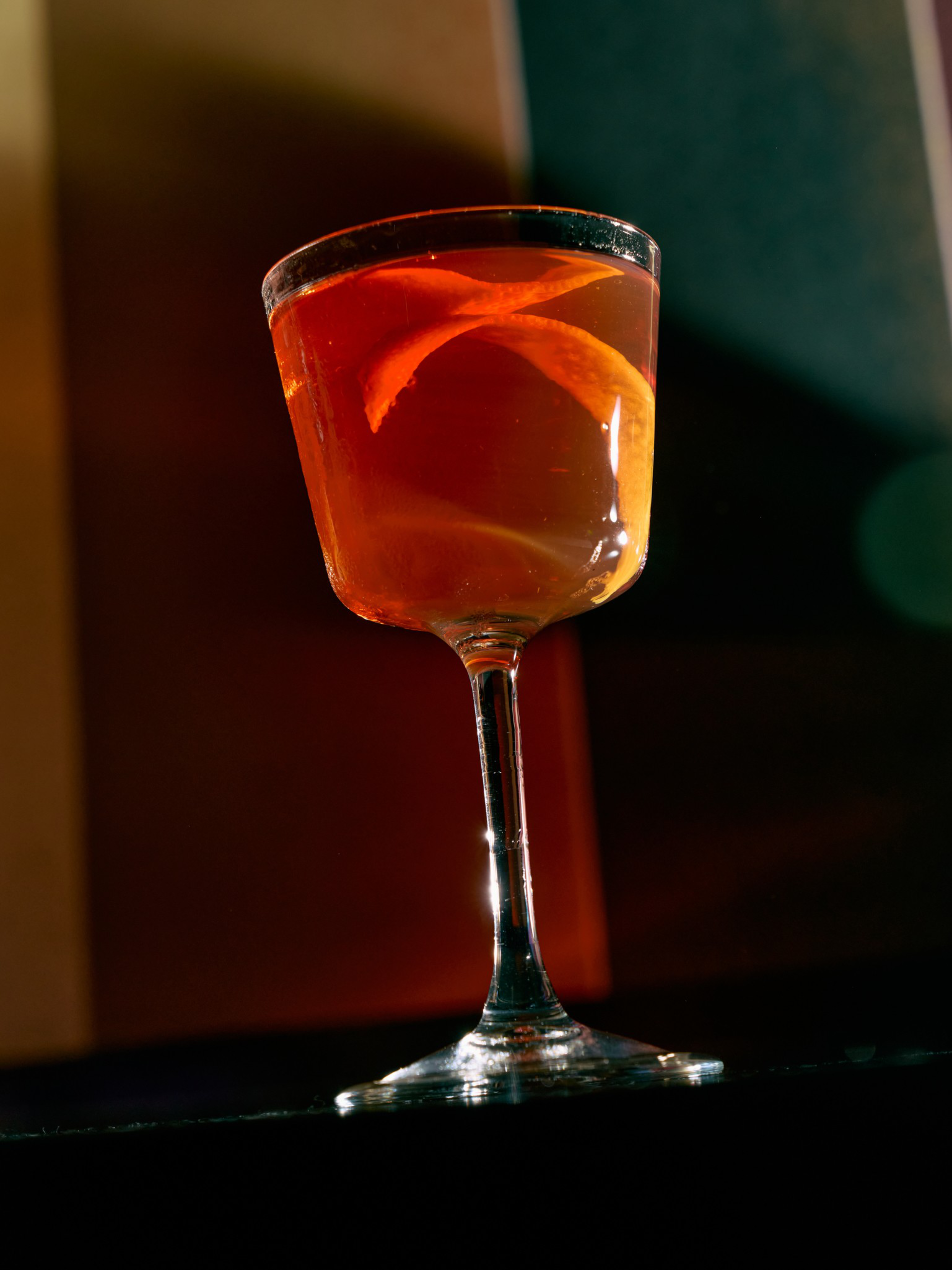 A stemmed glass contains an amber cocktail garnished with a twisted orange peel, set against a dark, softly lit background.