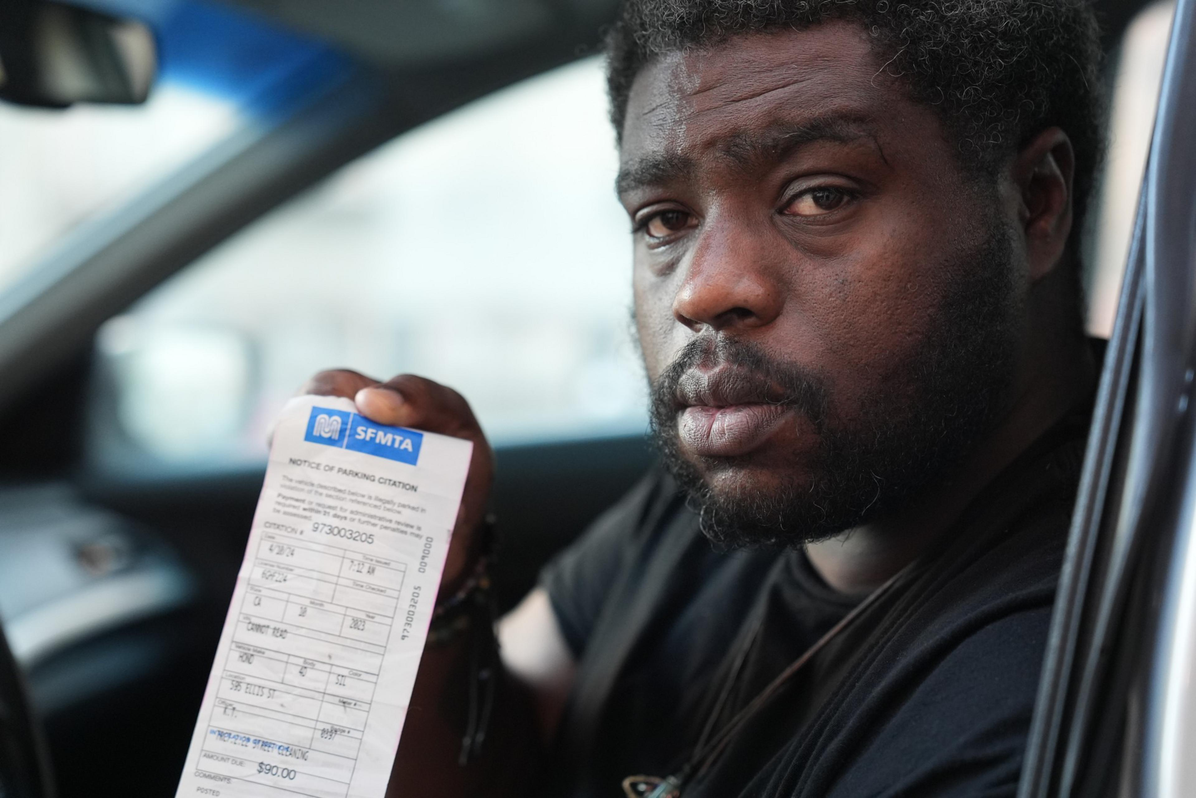 A man in a car, looking frustrated, holds up a parking citation.