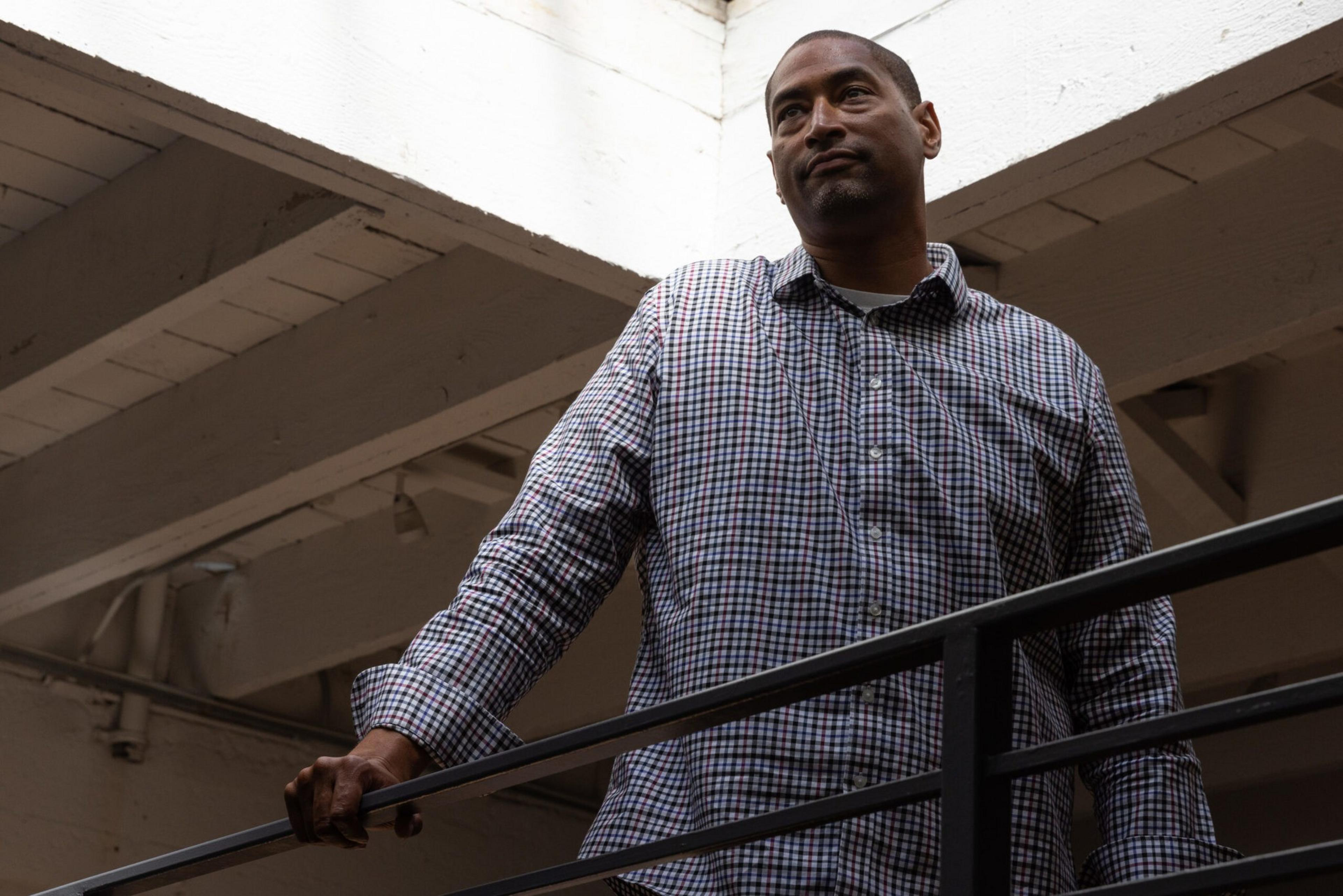 A man in a checkered shirt stands behind a railing, looking thoughtfully to the side in a dimly lit, industrial-style space with exposed beams.