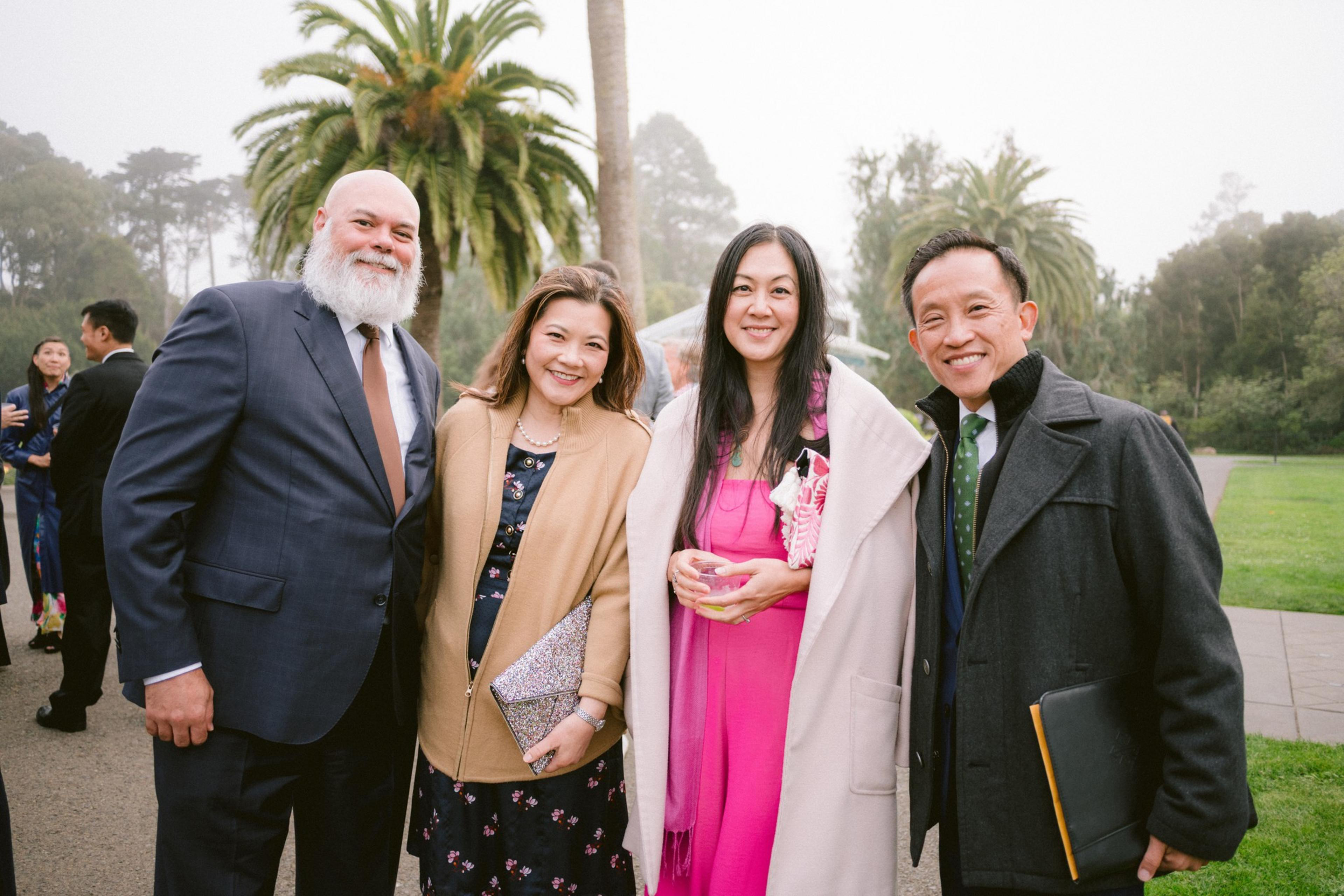 Four people stand close together outdoors, smiling; two women wear coats over dresses, one pink and one floral, while two men wear suits and ties.