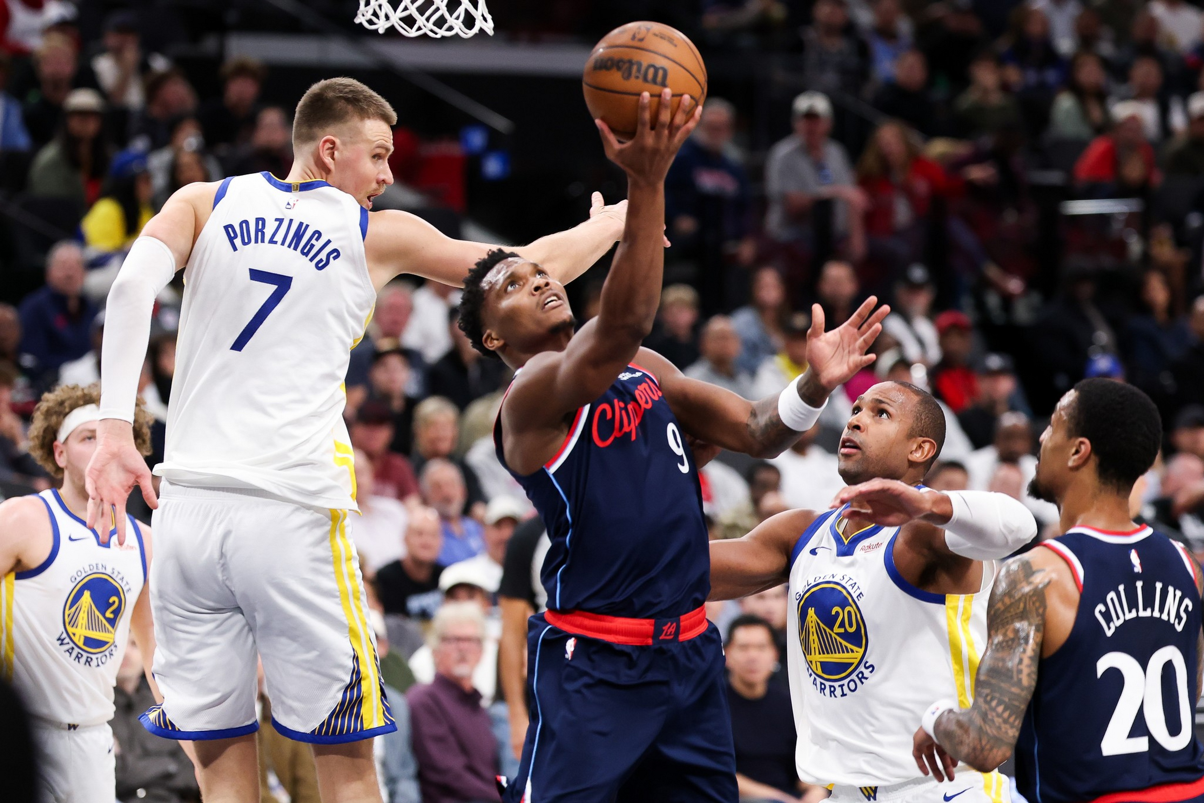 A Clippers player leaps to shoot while three Warriors players, including one with jersey number 7 and another number 20, attempt to block him.