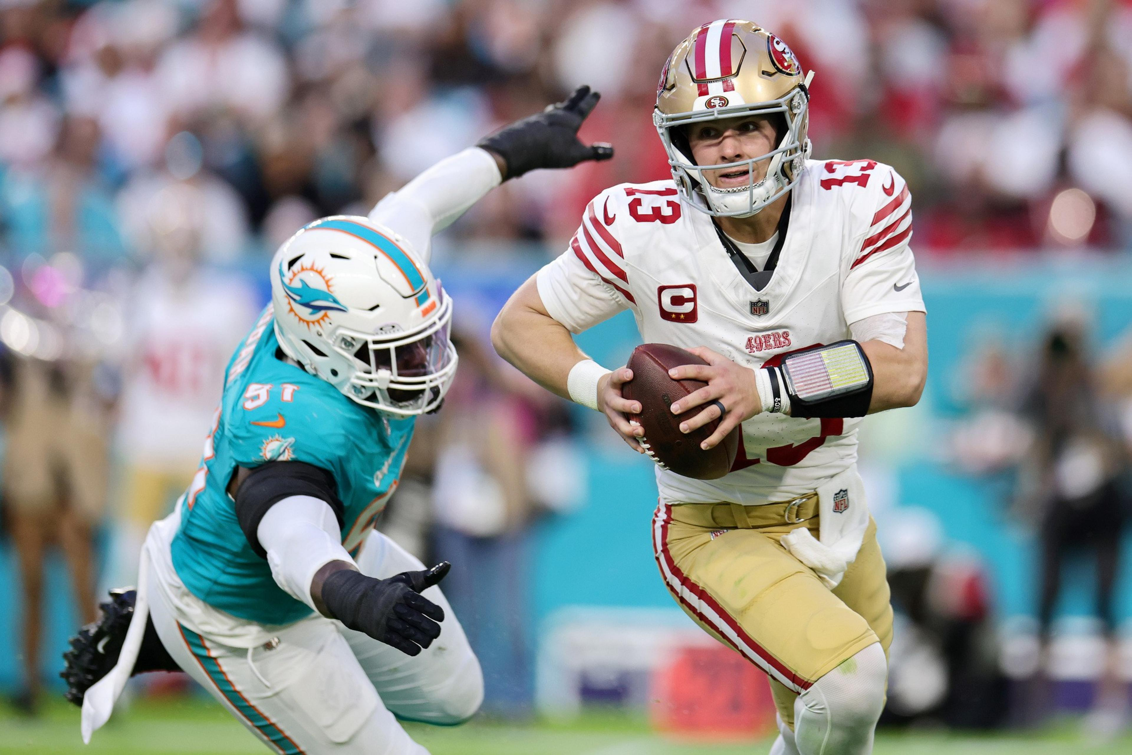 A football player in a red and gold uniform runs while holding the ball, pursued by a player in a turquoise and white uniform on a football field.