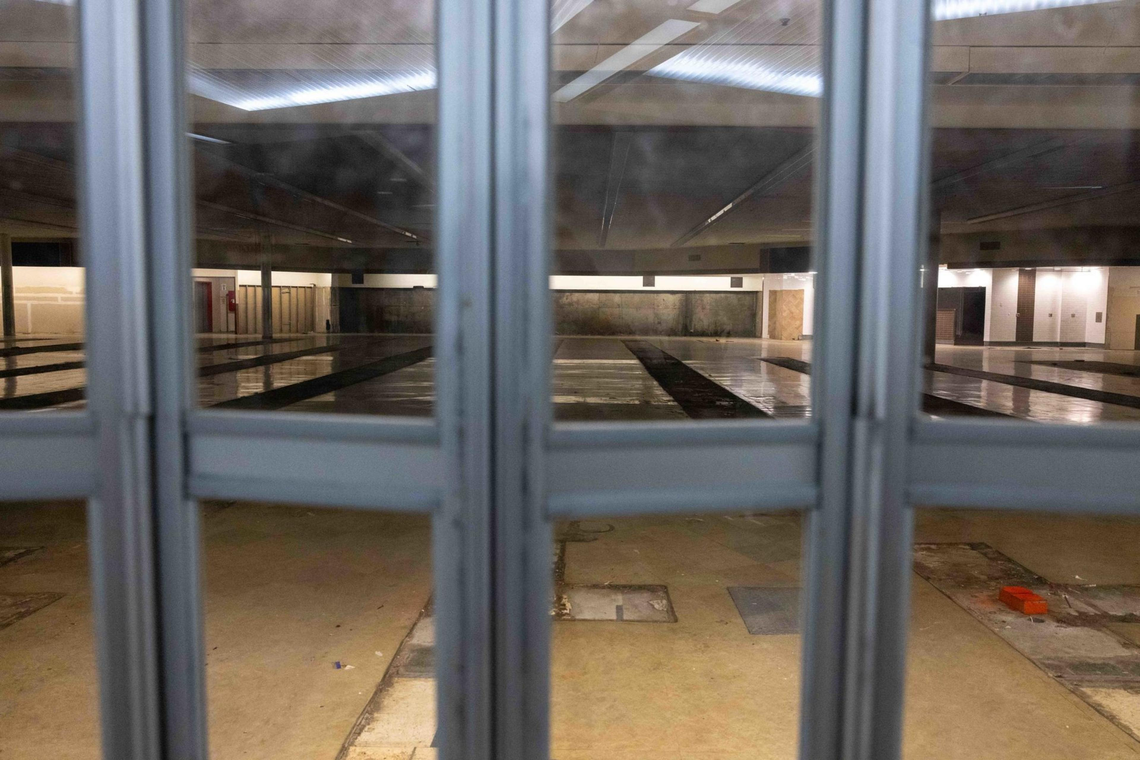 The image shows an empty indoor swimming pool, viewed through a set of glass doors, with lanes visible on the pool floor and overhead lighting.
